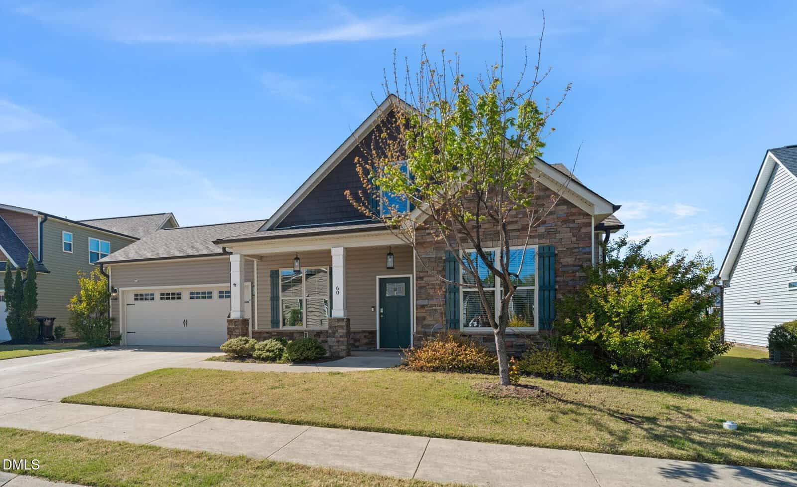 Single-story house with stone and siding exterior, two-car garage, covered front porch, and a small tree and bushes in the front yard. The house sits on a neatly maintained lawn bordered by a sidewalk.