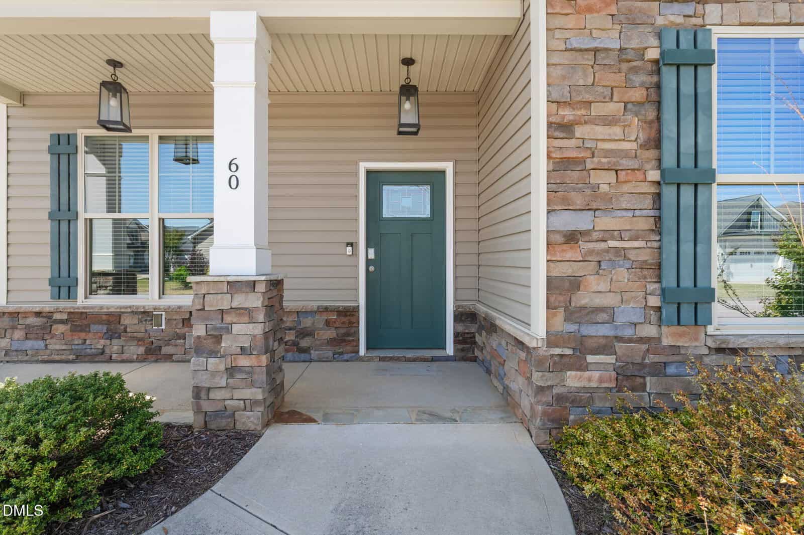Front entrance of a house with a green door, stone and siding exterior, white columns, outdoor lights, window shutters, and landscaped bushes along the walkway. The number 60 is on the column by the front door.