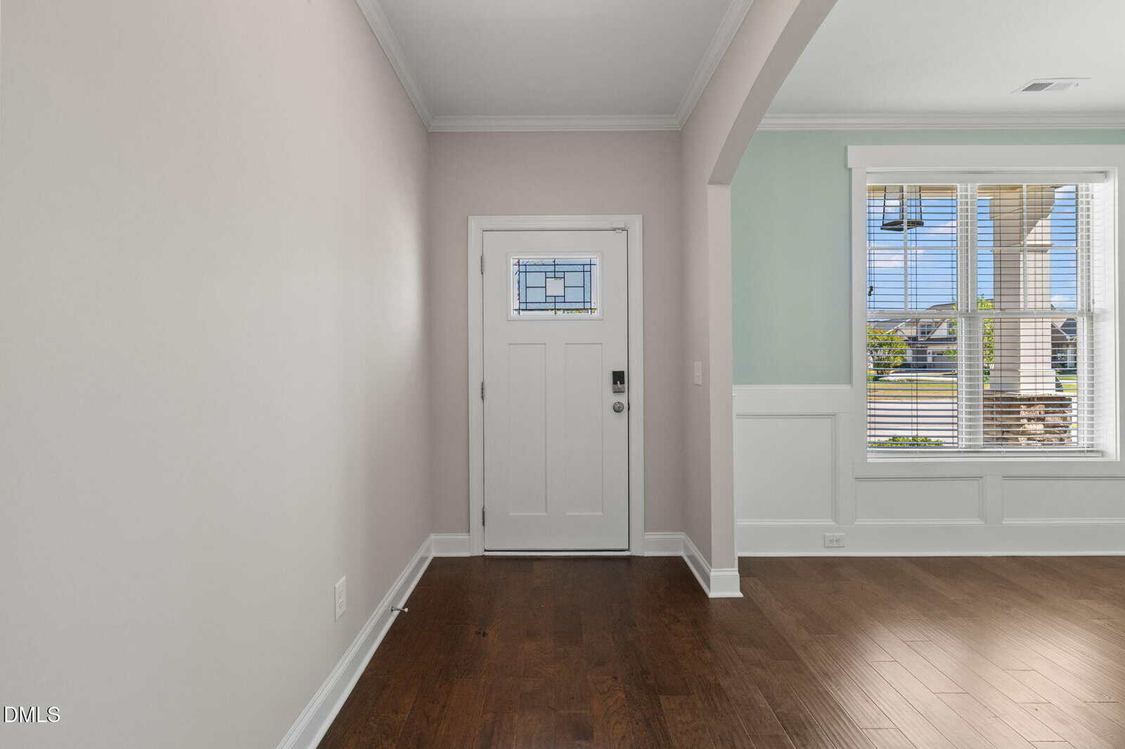 A white front door with a small window and silver hardware is set in a light-colored wall. Next to it, a window with white trim lets in sunlight. The floor is dark wood, and an archway separates the entry from another room.