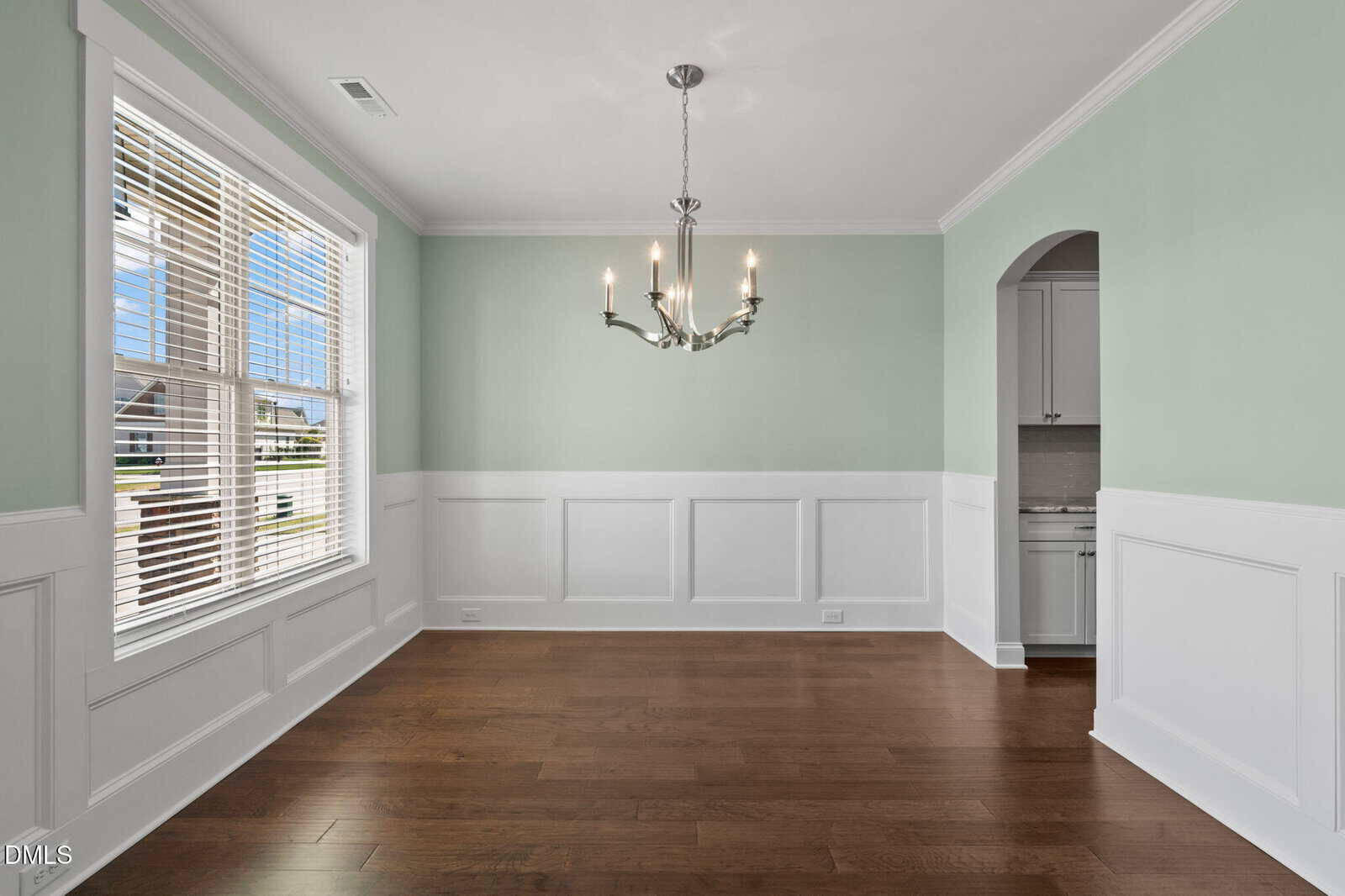 Empty dining room with light green and white paneled walls, a metal chandelier, large window with blinds, and brown wood floors. An arched doorway leads to an adjacent room with white cabinetry.