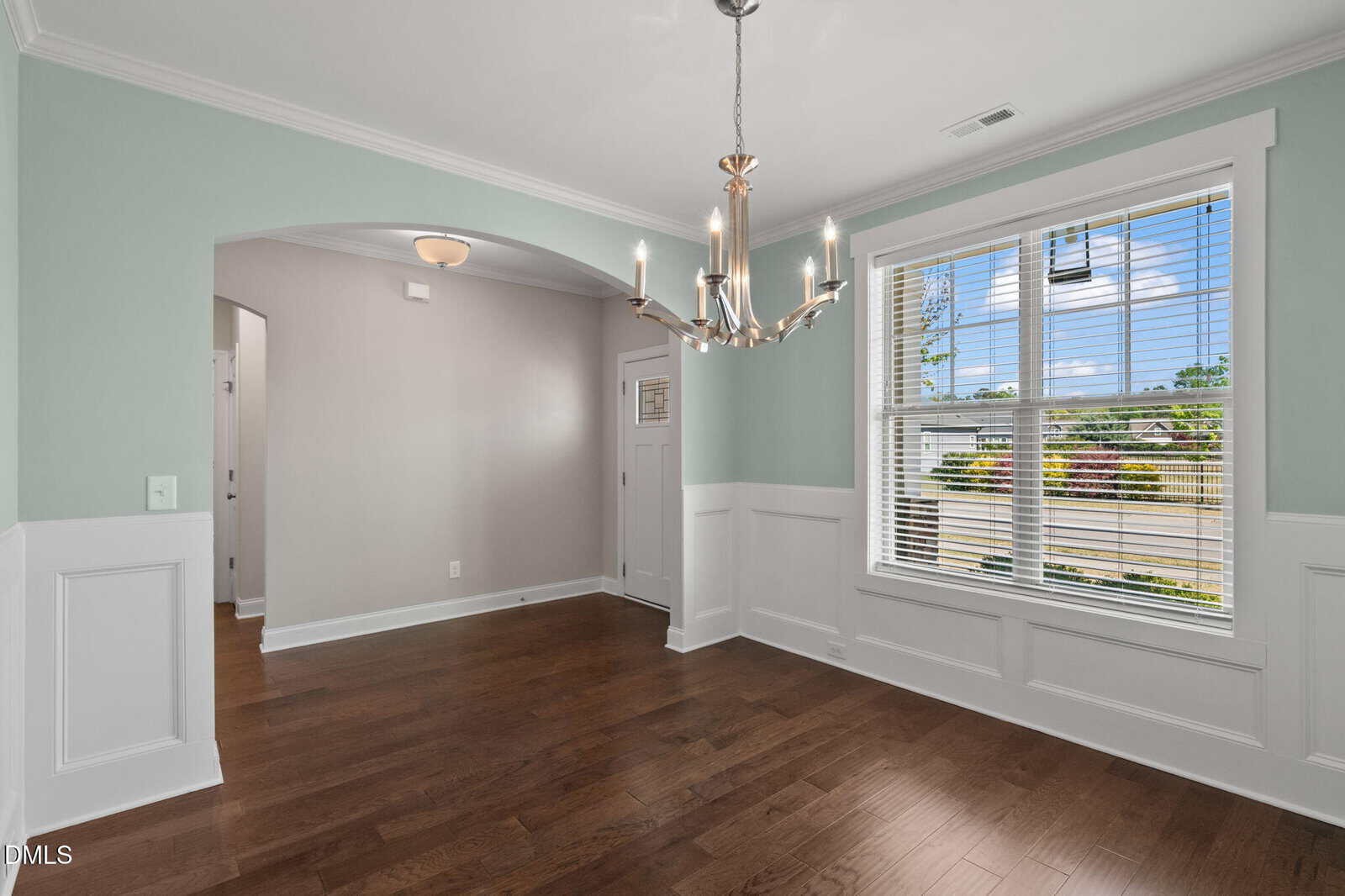 A bright dining room with hardwood floors, soft green and white walls, a modern chandelier, large window with blinds, and an arched entryway leading to another part of the house.