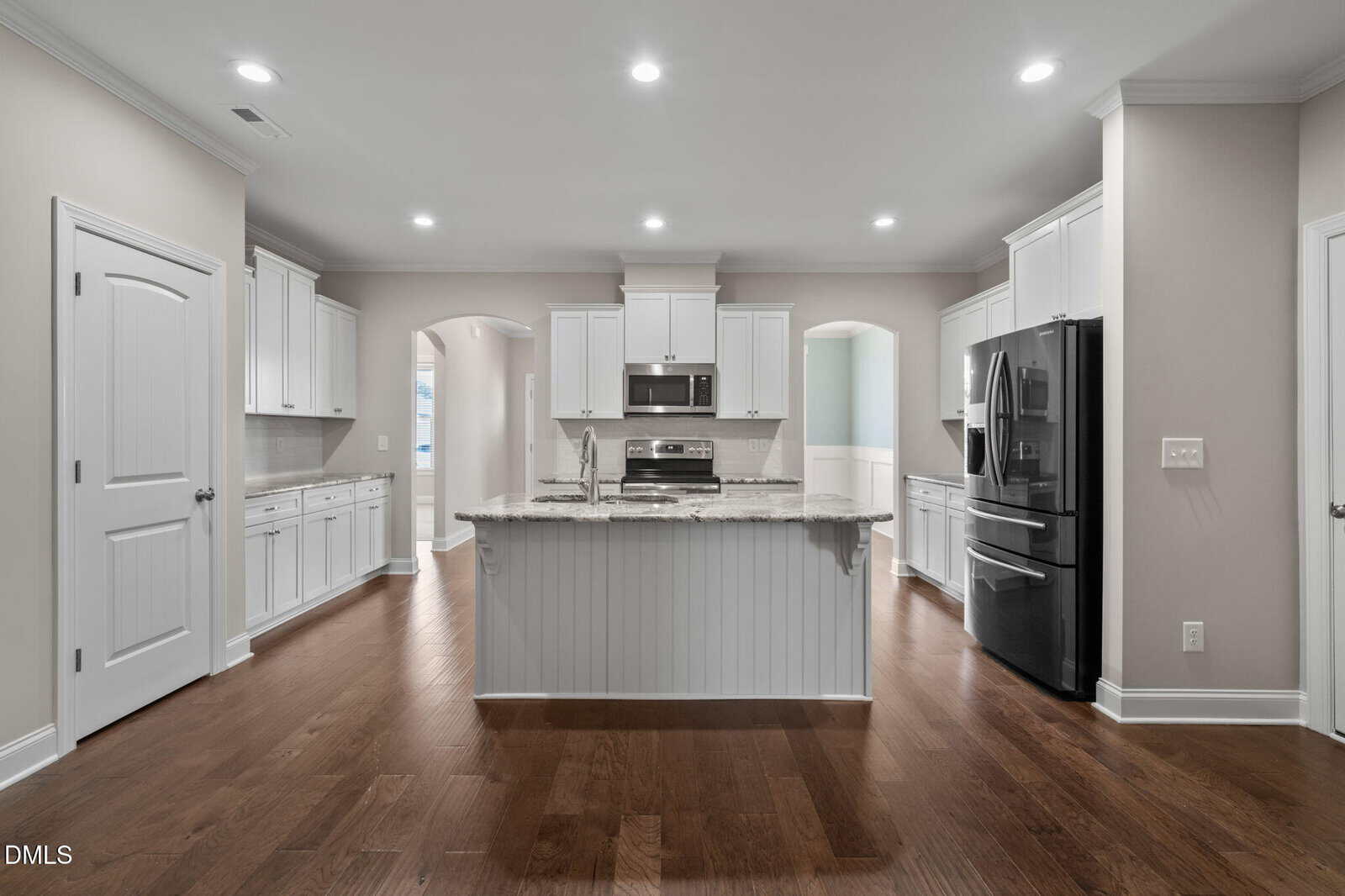 Modern kitchen with white cabinets, stainless steel appliances, a black refrigerator, granite island countertop, hardwood floors, and recessed lighting. Two arched doorways lead to other rooms.