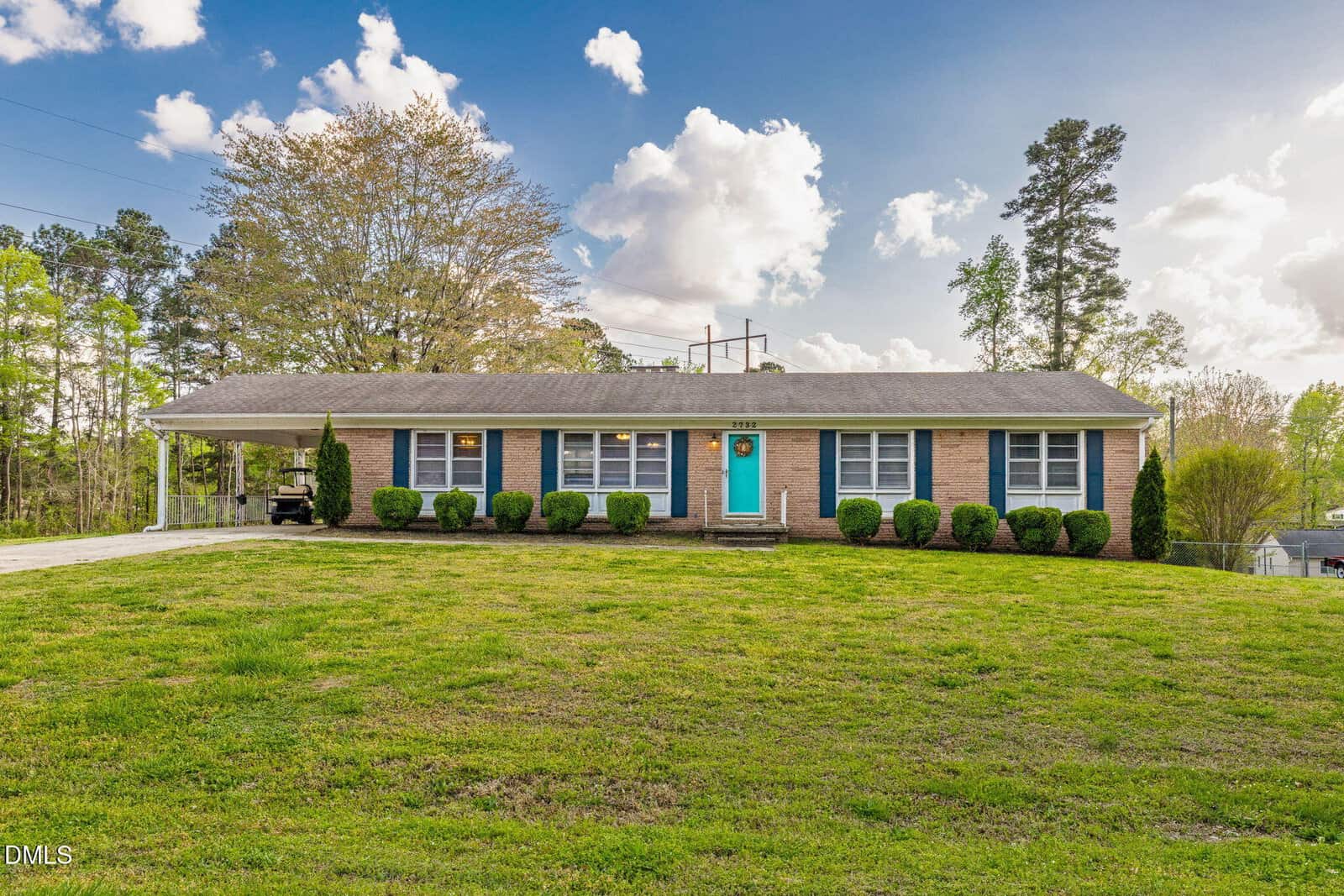 Single-story brick house with white trim, a bright blue front door, and eight front windows at 2732 Nutbush Road, Henderson. Trimmed bushes line the facade, and a large green lawn covers the front yard. Tall trees and a partly cloudy sky are in the background.