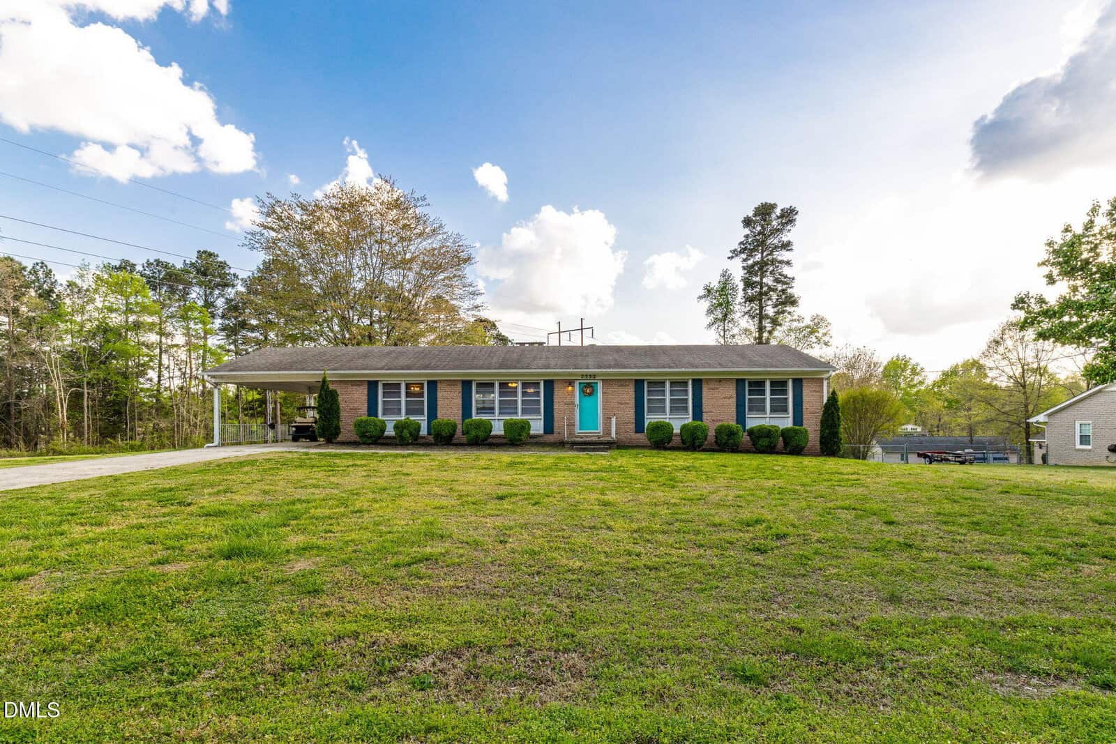 Single-story brick house with blue shutters, a central blue door, and attached carport, situated on a large grassy lawn with trees and a cloudy sky in the background.