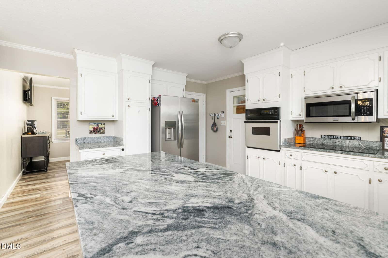 Modern kitchen with white cabinets, stainless steel appliances, a gray marbled countertop island, light wood flooring, and neutral walls. A small kitchen nook with a window and TV is visible through an open doorway.