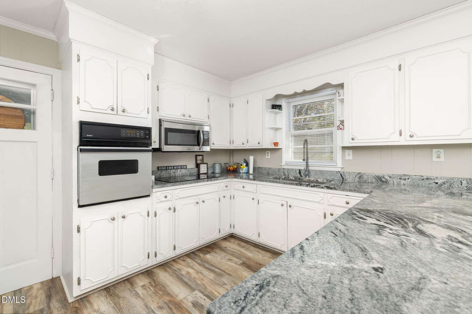 A kitchen with white cabinets, stainless steel appliances, a double oven, a microwave, light gray marble countertops, wood-style flooring, a sink under a window, and small decorative items on the counters and shelves.