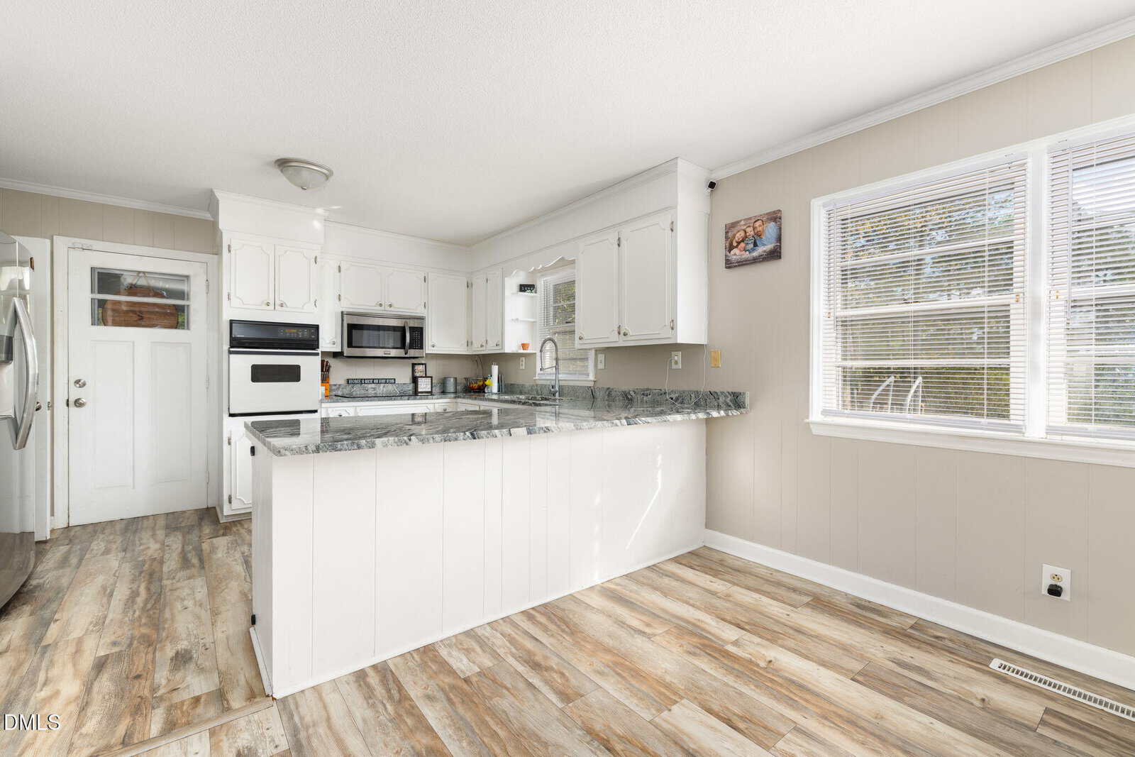 Bright kitchen with white cabinets, marble countertops, stainless steel appliances, light wood flooring, and a large window on the right letting in natural light. There is a door and built-in oven on the left wall.