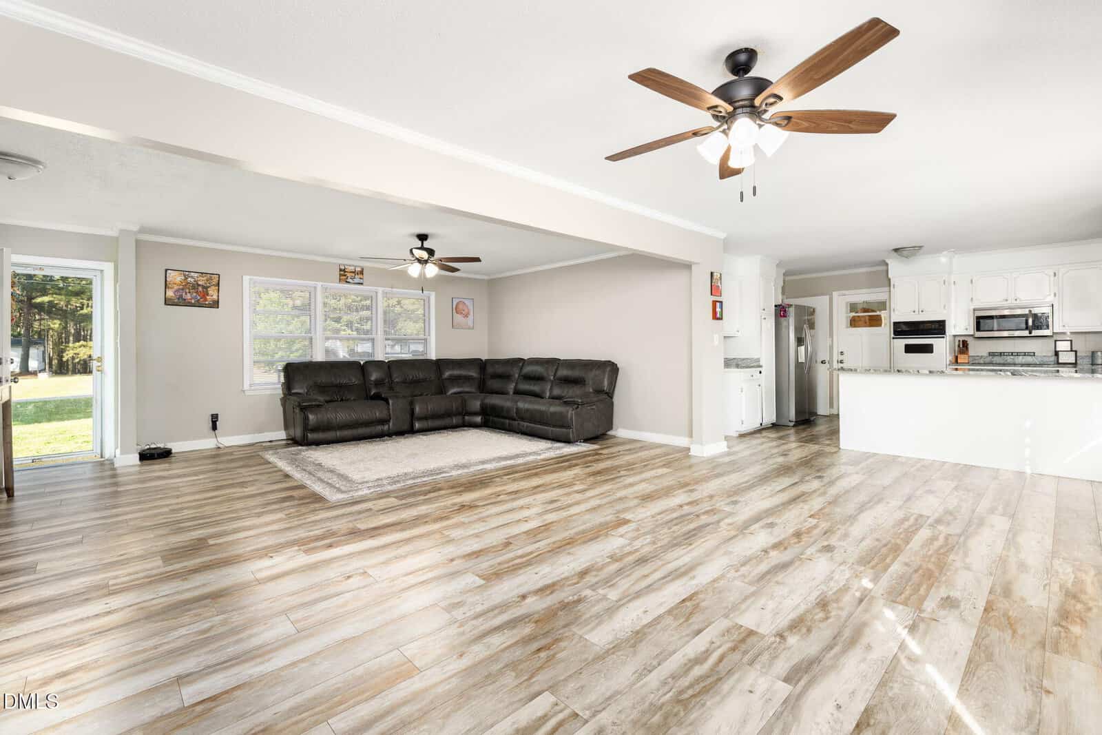 Spacious open-concept living room and kitchen with light wood flooring, two ceiling fans, a large brown sectional sofa, and white cabinets. Sunlight streams in from windows and a glass door.