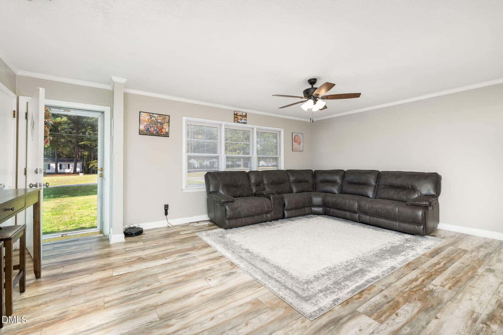 A bright living room with wood-look flooring, a large dark brown sectional sofa, a light area rug, two windows, an open door, a small table, and ceiling fan with lights. Art decorates the light-colored walls.