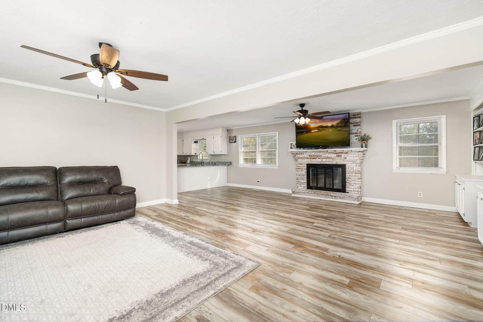 Spacious living room with wood-style flooring, ceiling fans, a dark sectional sofa, a light area rug, a brick fireplace with a TV above it, built-in shelves, and large windows providing natural light.