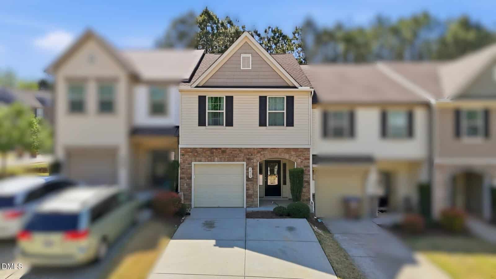 A two-story townhouse with beige siding, brick accents, a single garage, and a driveway, flanked by similar houses; bushes and a small tree line the front entry.