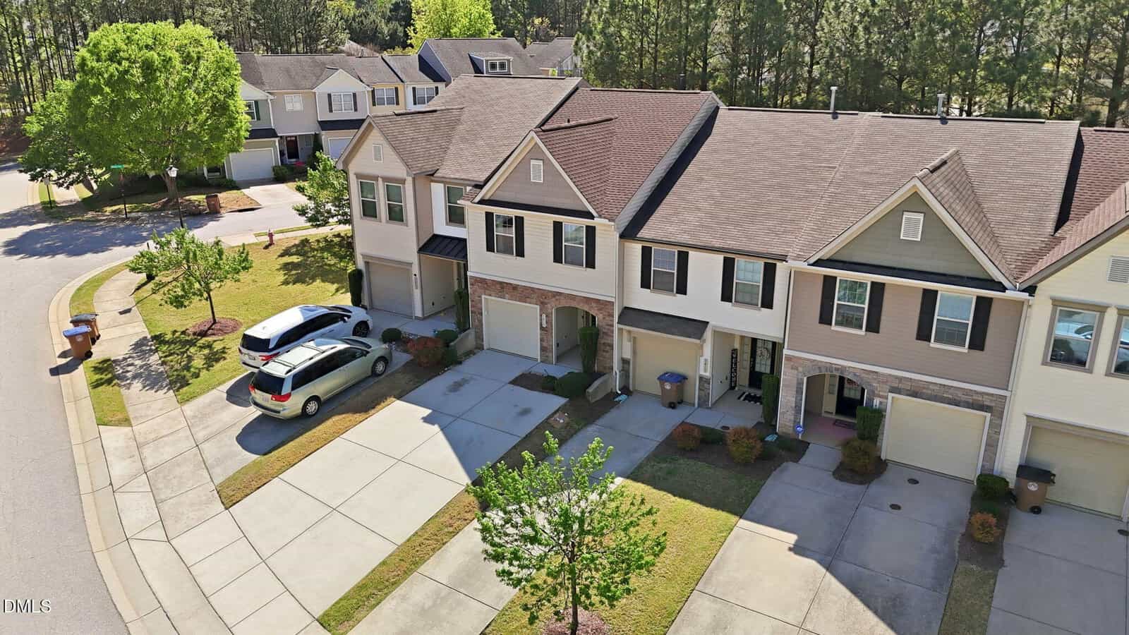 A row of modern two-story townhouses with attached garages and driveways, cars parked outside, and neatly maintained lawns along a curved suburban street with trees and sidewalks.