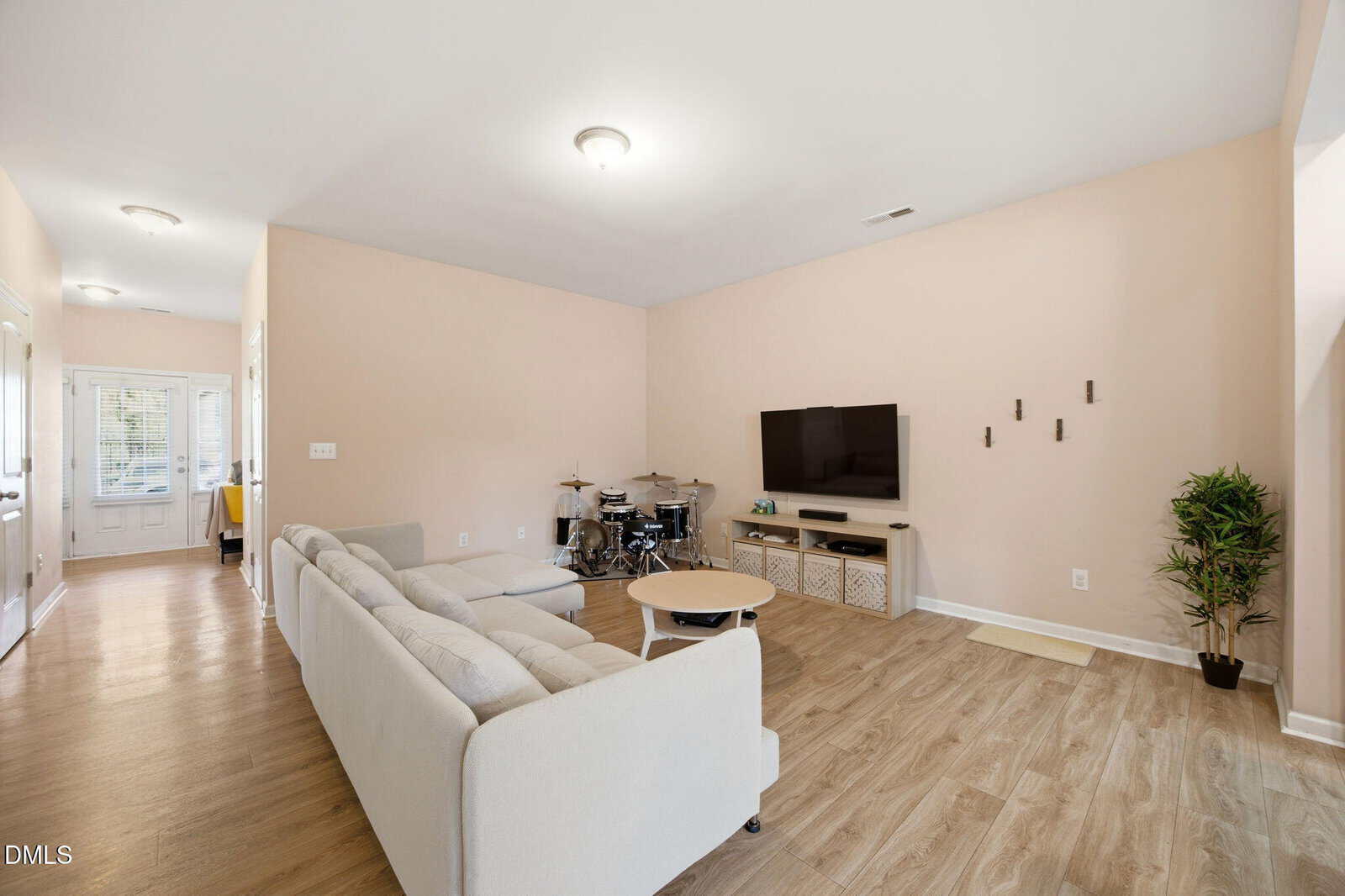 A bright, modern living room with light wood flooring, beige walls, a light-colored sectional sofa, a round coffee table, a wall-mounted TV, a small drum set, and a potted plant in the corner.