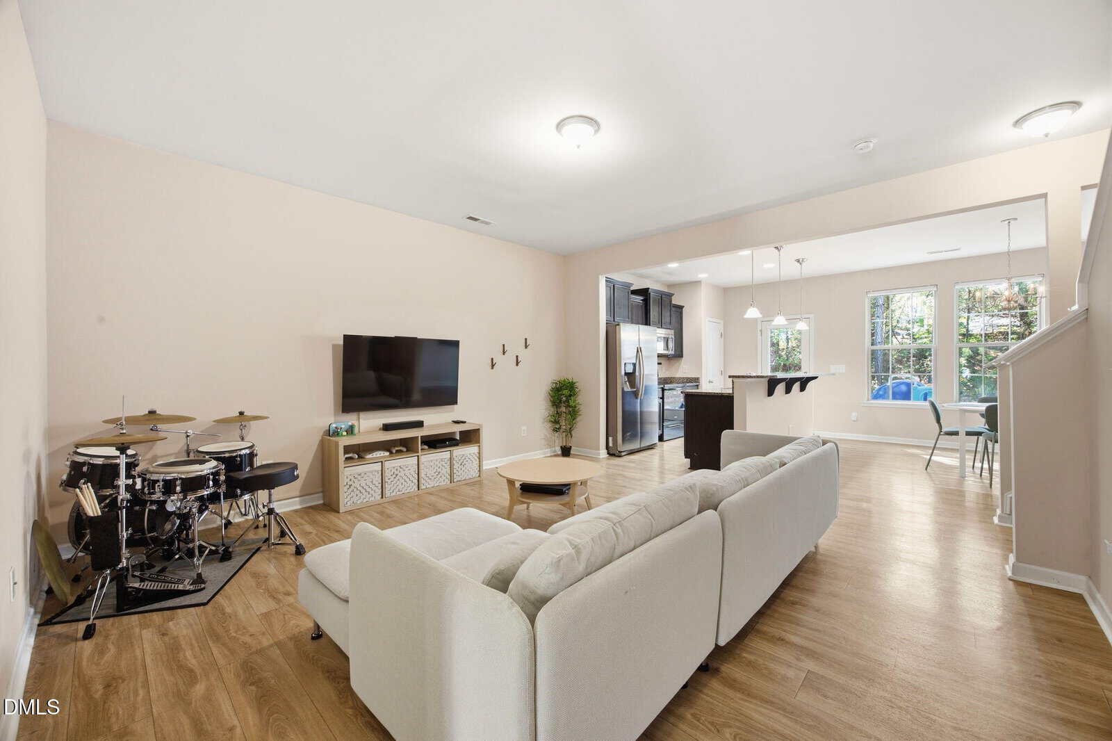 Open living room with light wood floors, a beige sectional sofa, wall-mounted TV, electronic drum set, and an open kitchen and dining area with large windows in the background.