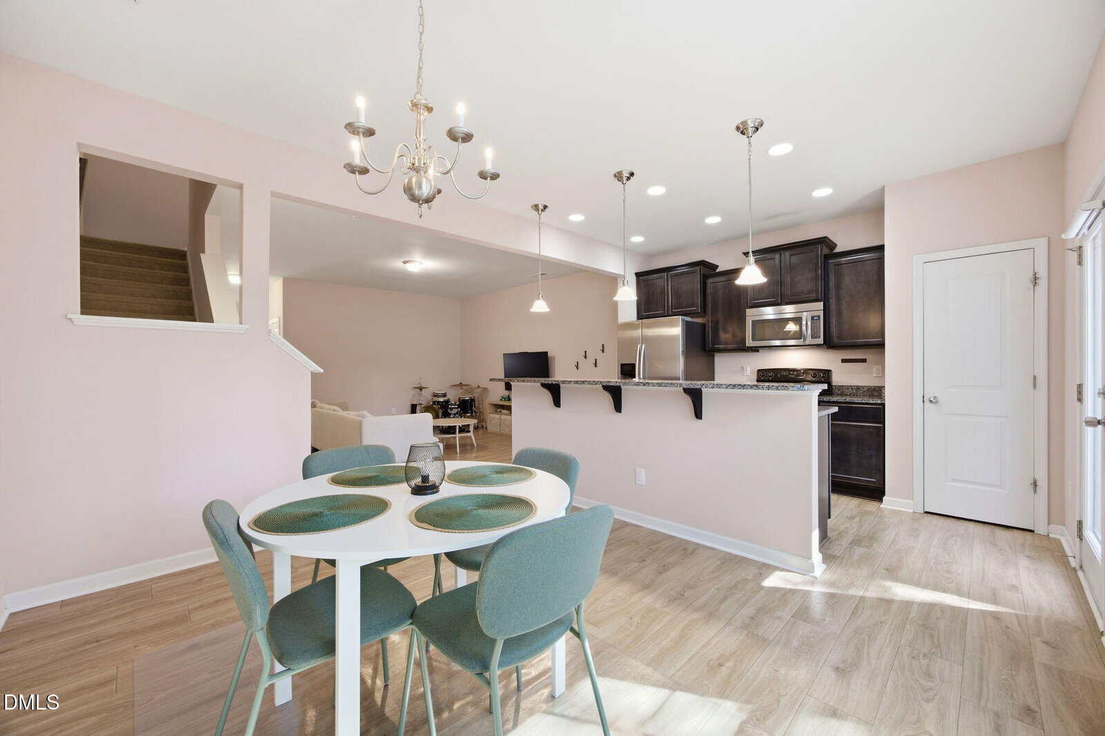 Open-concept kitchen and dining area with pastel walls, wood flooring, round dining table set for four, modern bar stools, stainless steel appliances, and a cozy living space in the background. Natural light fills the room.