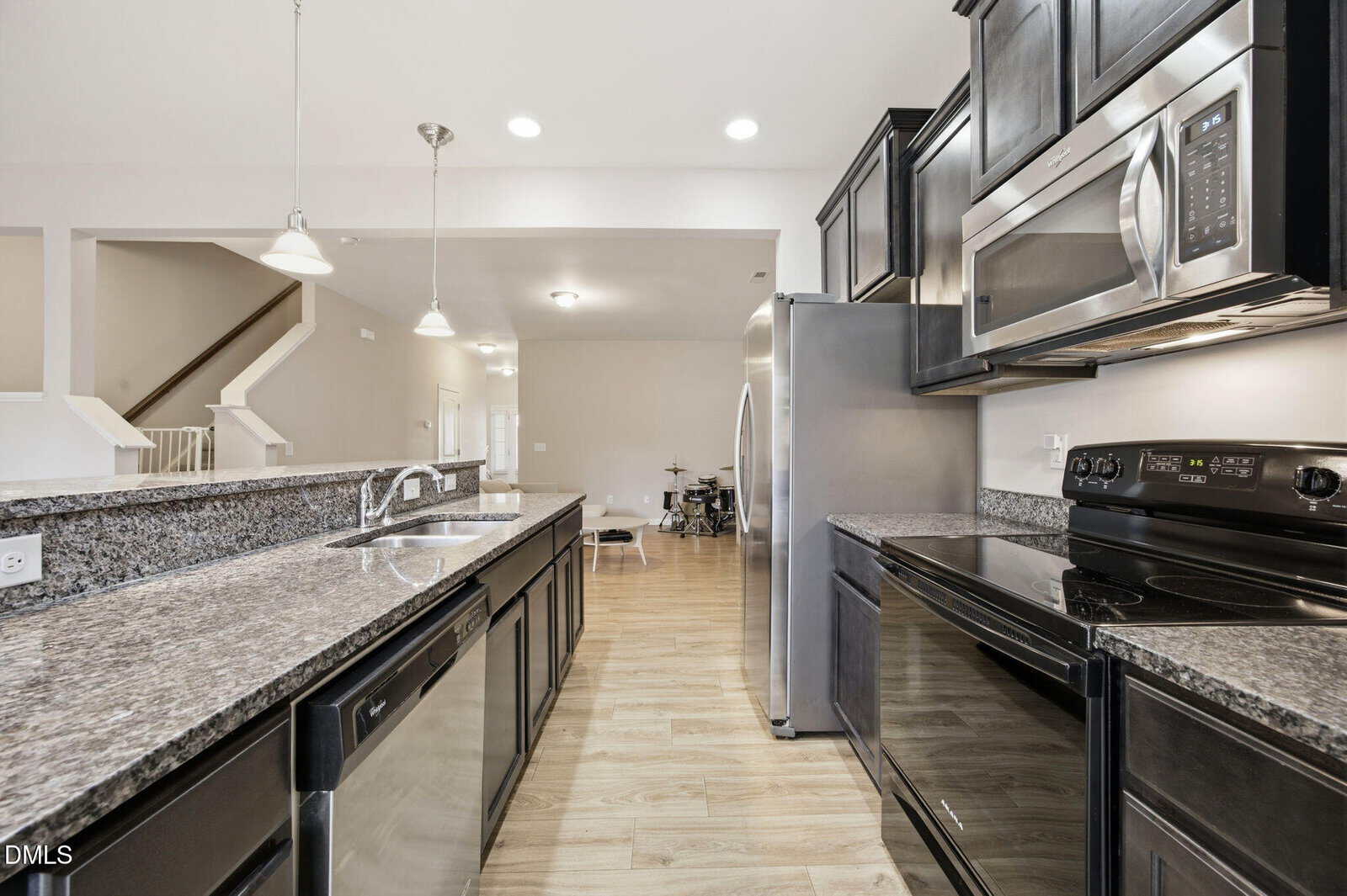 Modern kitchen with dark cabinets, stainless steel appliances, granite countertops, and light wood flooring. The space opens to a living area with visible stairs and a dining nook in the background.