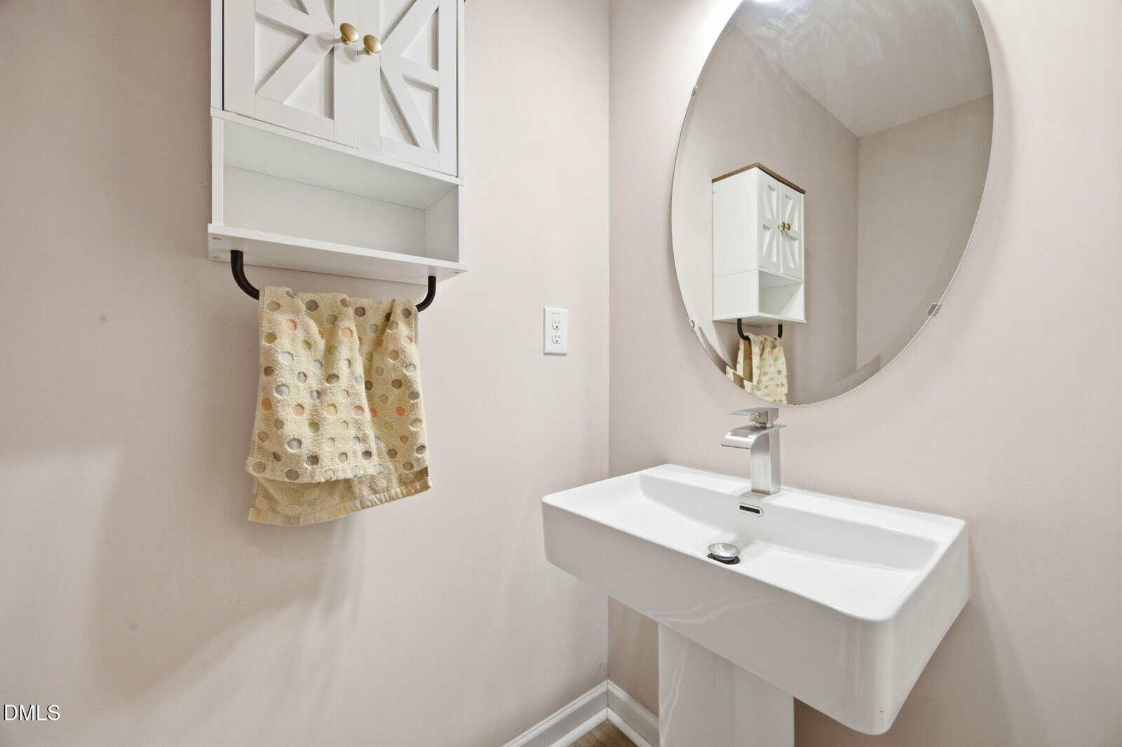 A modern bathroom with a white pedestal sink, a round mirror above it, a wall-mounted cabinet, and a yellow patterned hand towel hanging below the cabinet. The walls are light-colored.