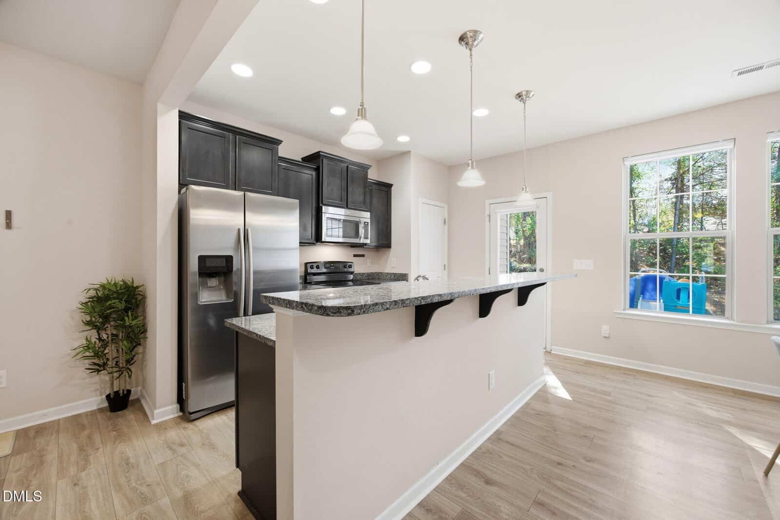 Modern kitchen with black cabinets, stainless steel appliances, granite countertops, pendant lighting, and a breakfast bar. Large windows let in natural light, and there is a small indoor plant in the corner.