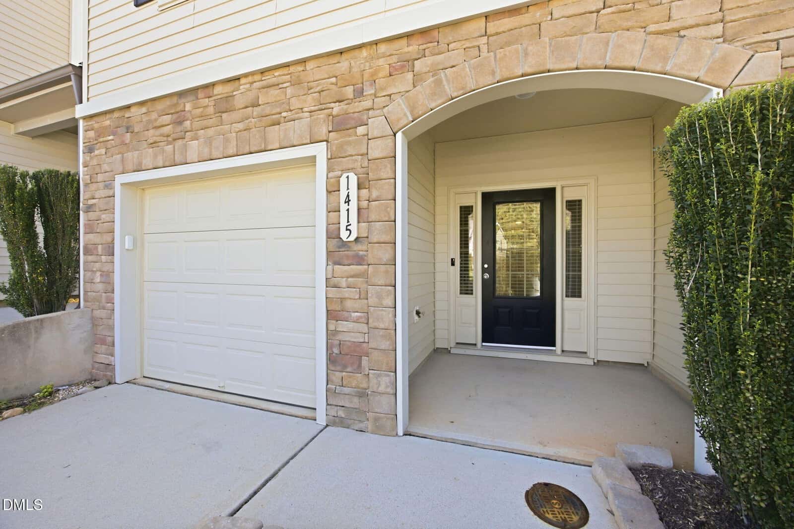 A house exterior with tan stone and siding, a white garage door, the address number 1415, an arched entryway, neatly trimmed shrubs, and a black front door with side windows.