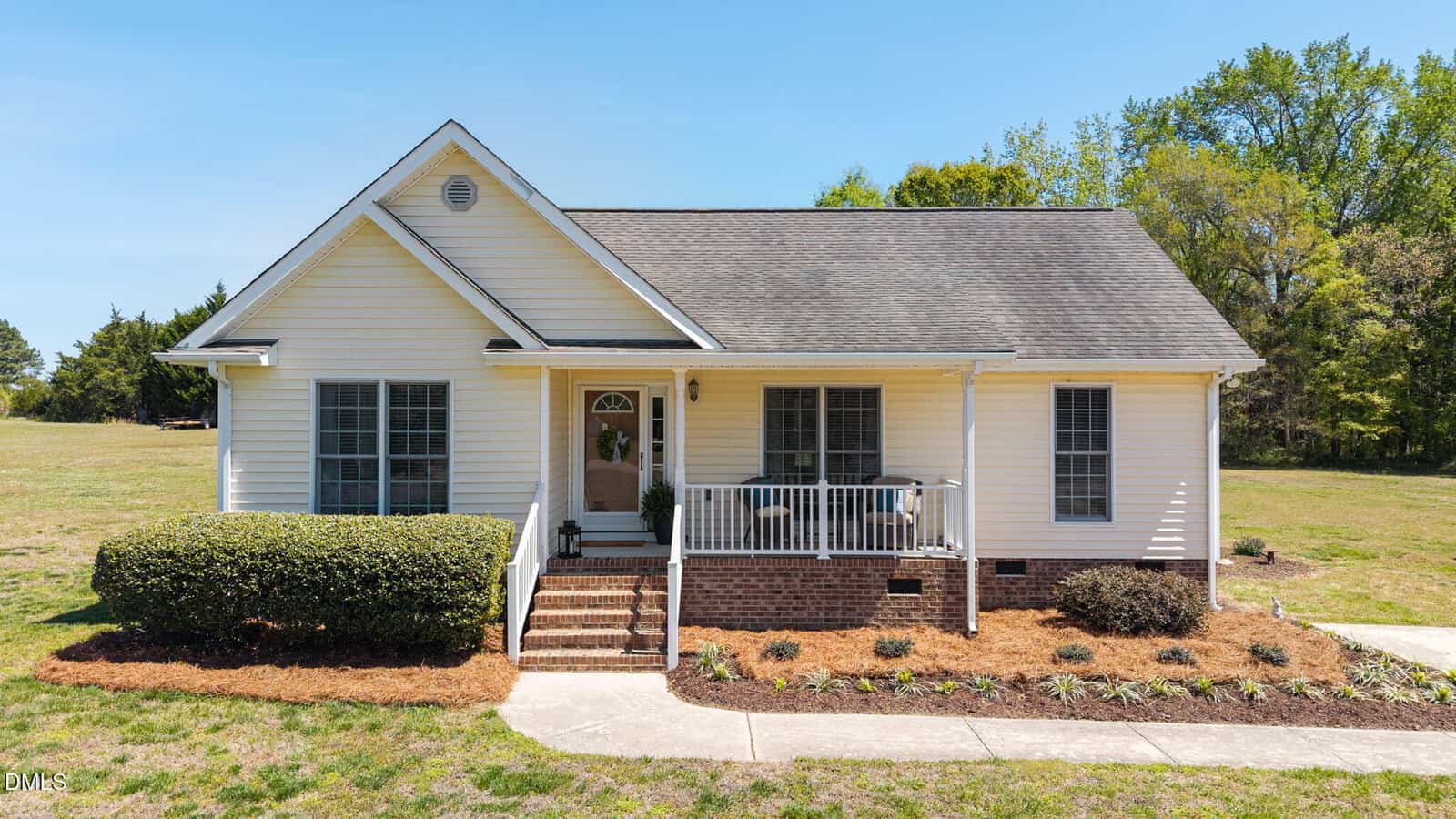 A small white house with a front porch, steps, and railing sits gracefully at 65 Candlewood Drive in Zebulon. It features a brick foundation, neatly trimmed bushes, and landscaped garden beds in the front. The yard is grassy, with trees and a clear blue sky in the background.