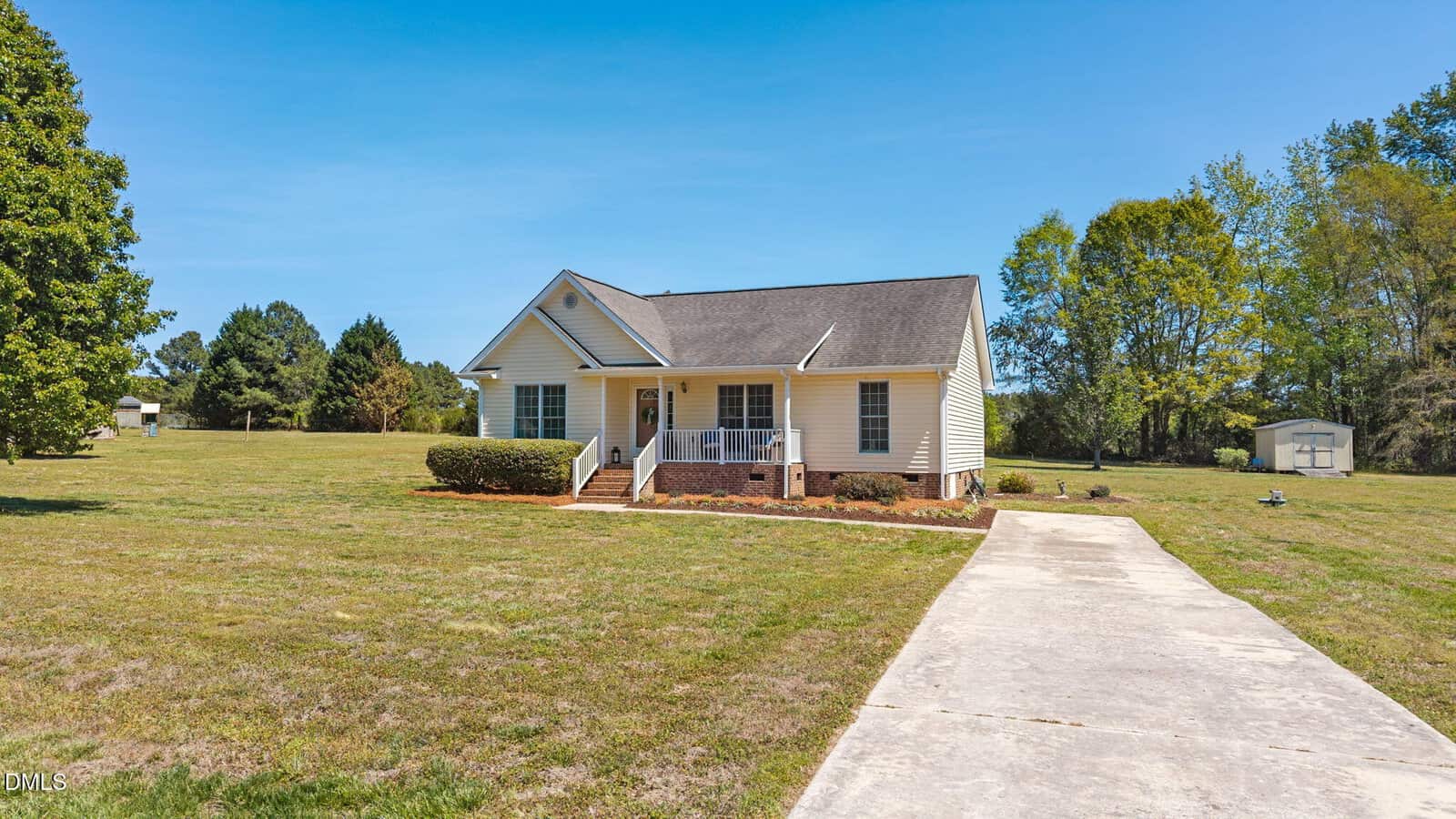 Single-story yellow house with a front porch, brick steps, and white trim, set on a large grassy lot with trees in the background and a long concrete driveway leading to the entrance.