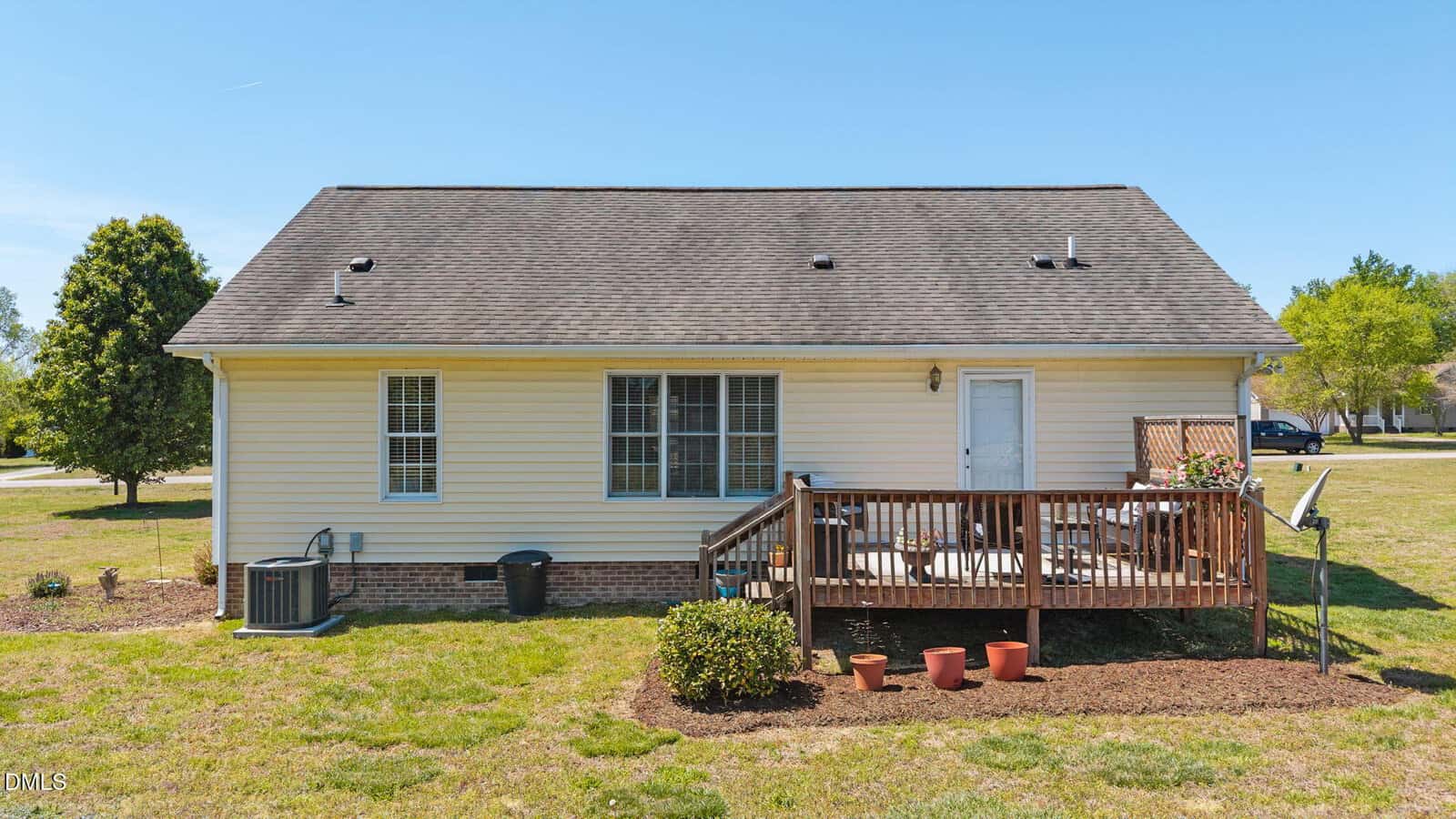 A single-story beige house with a shingled roof, small wooden deck, outdoor seating, and several potted plants in the backyard, surrounded by grass and a few trees on a sunny day.