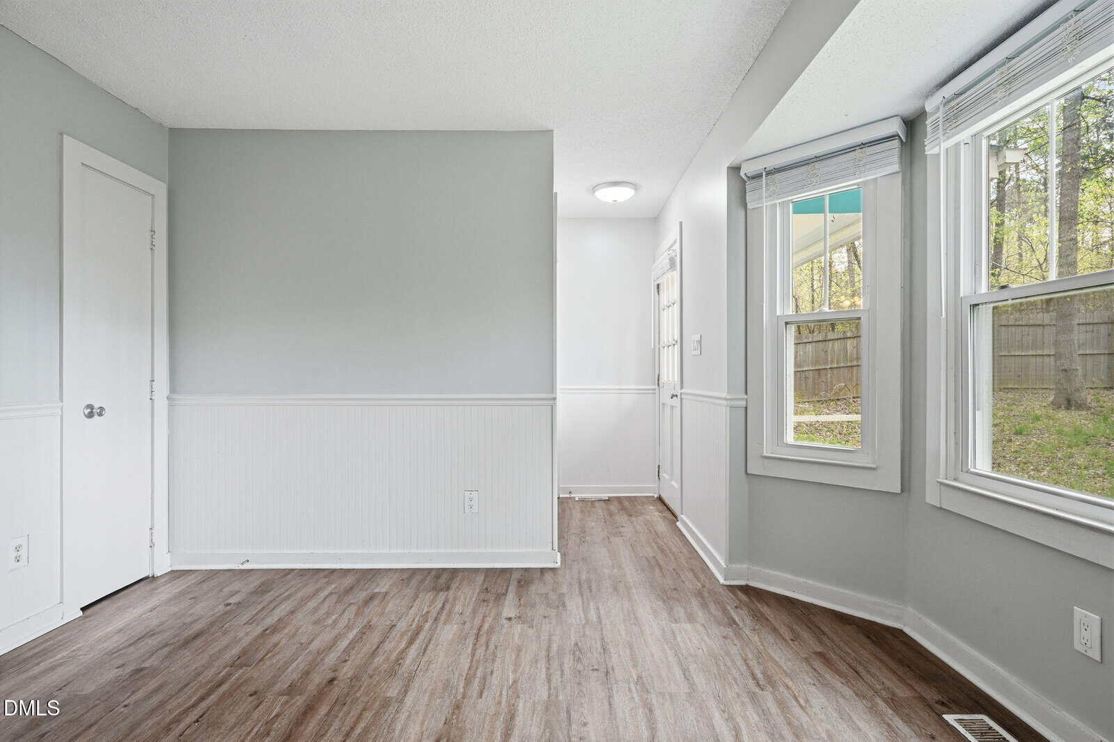 A small, empty room with light gray walls, white trim, wood-look flooring, two windows with blinds, a door in the back corner, and a closed door on the left wall. Outside, a fenced yard is visible through the windows.