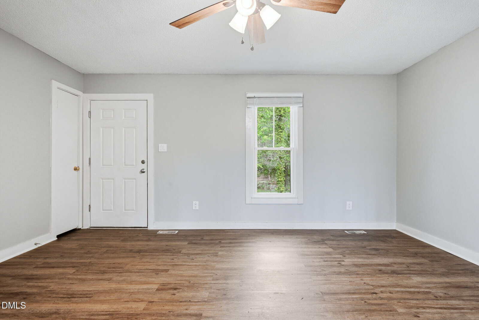 Empty room with light gray walls, a single window, a white door, wood-style flooring, and a ceiling fan with lights. No furniture or decorations are present.