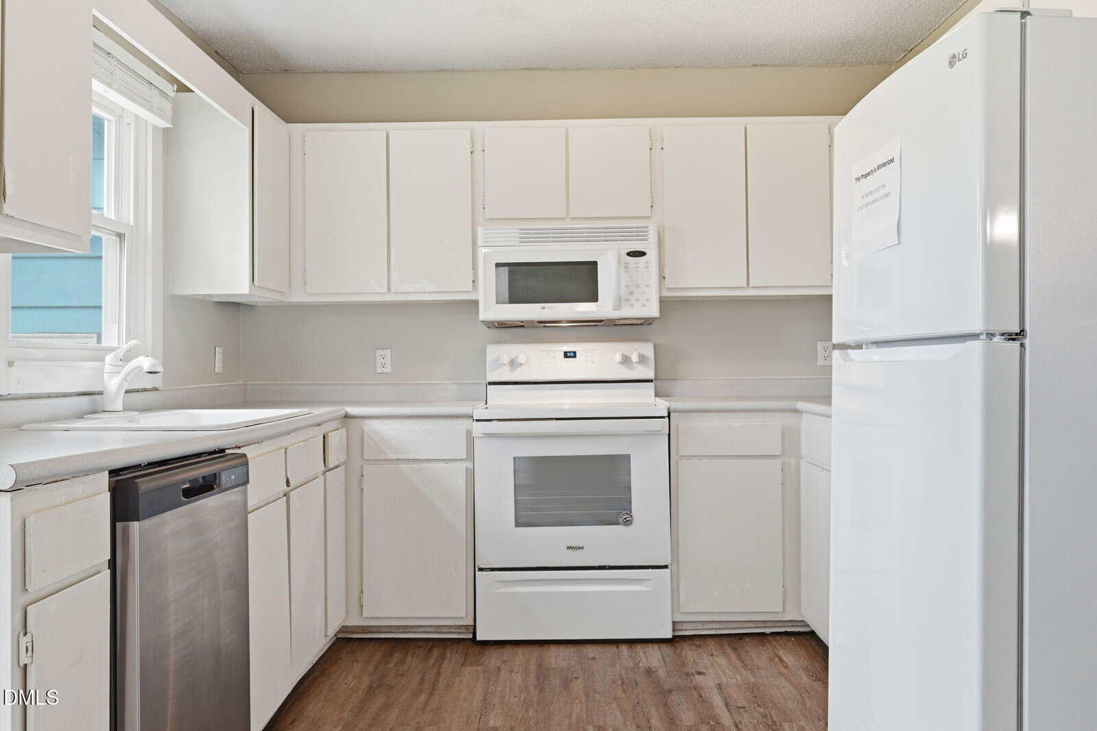A kitchen with white cabinets, white refrigerator, microwave, and oven. A stainless steel dishwasher is next to a white sink under a window. The floor has wood-style flooring. The walls are light-colored.