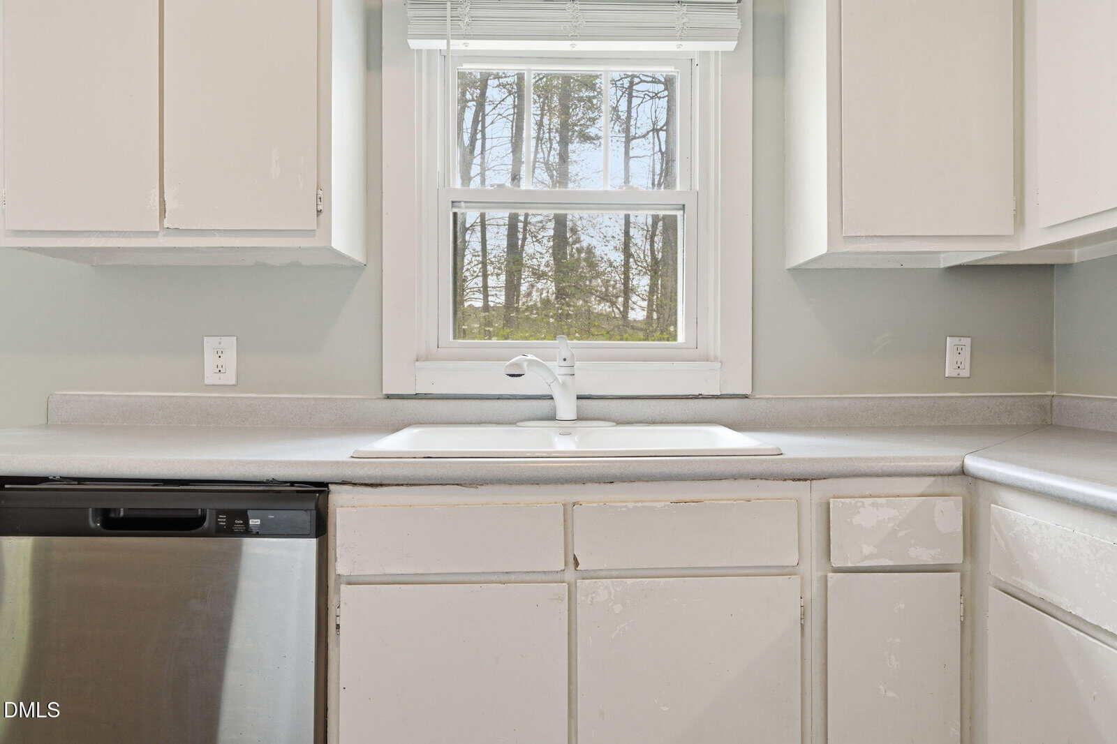 A white kitchen sink with a single faucet is set in a light counter beneath a window with blinds. The cabinets and drawers show worn paint. A dishwasher is on the left, and there are power outlets on the wall.