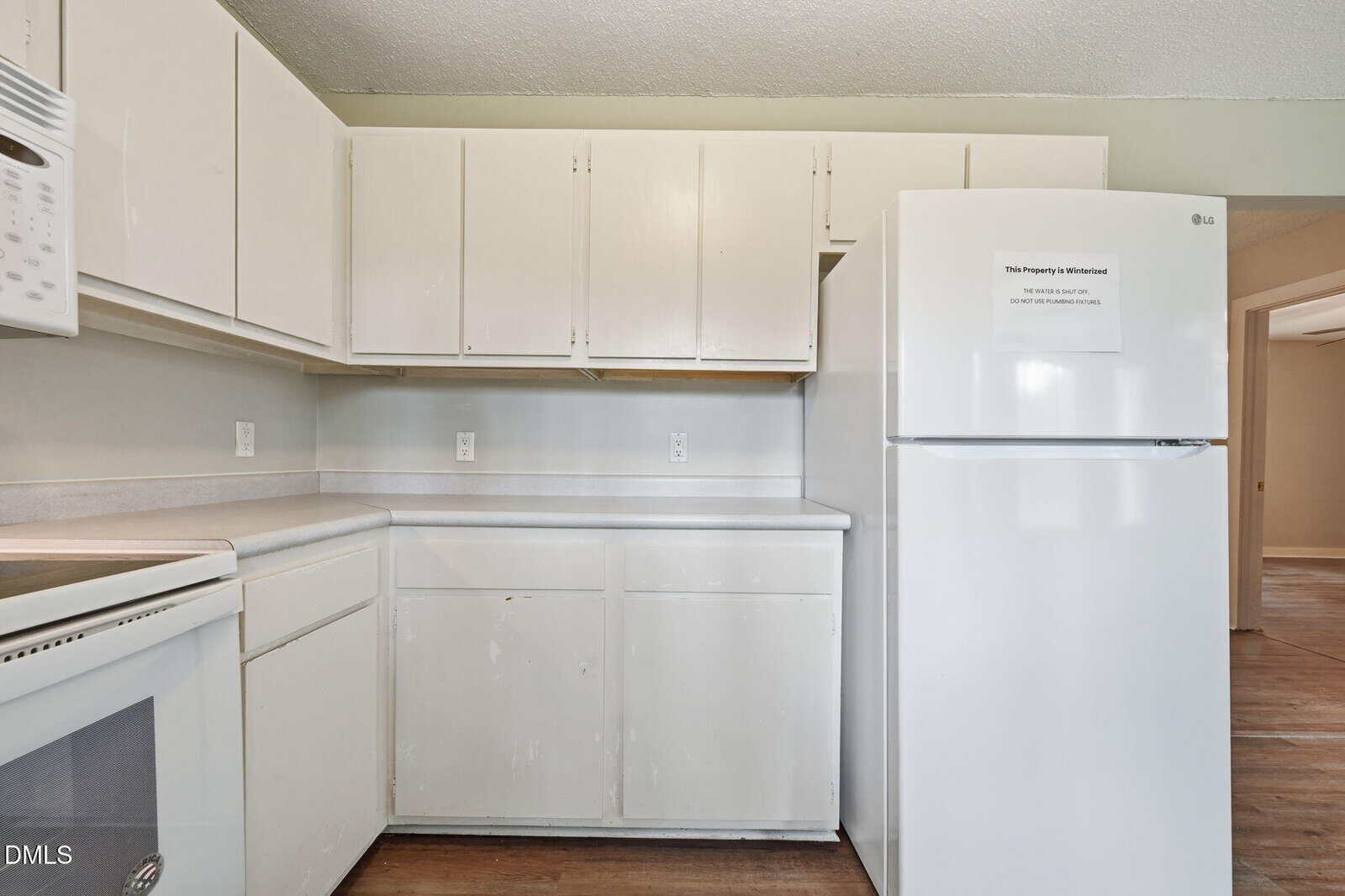 A kitchen with white cabinets, white appliances including a refrigerator and dishwasher, light countertops, and wood flooring. An open doorway leads to another room.