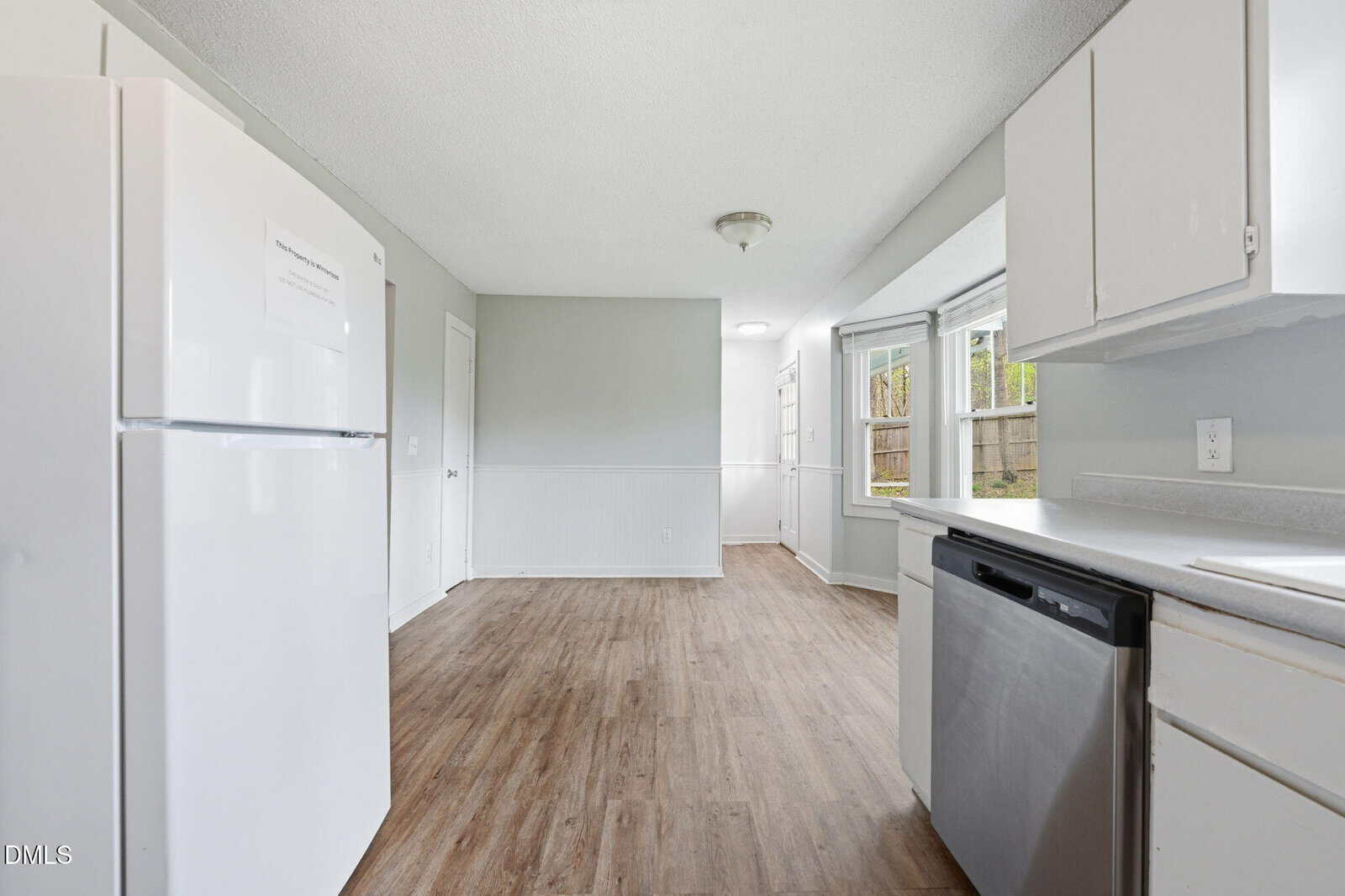 Bright kitchen with white cabinets, refrigerator, and dishwasher; wood flooring; large window letting in natural light; neutral wall colors; dining nook visible in the background; door and window on the far wall.