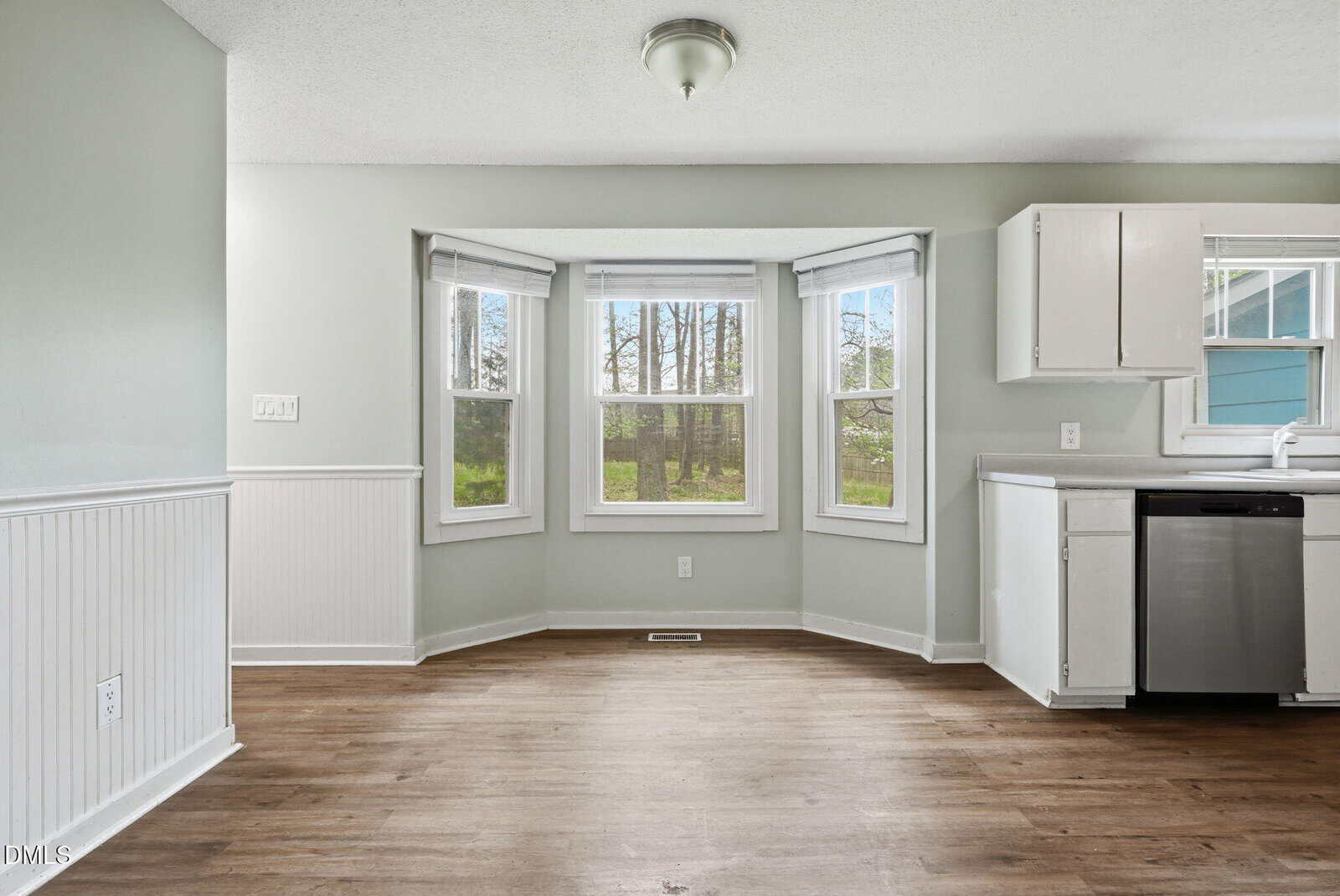A bright dining area with three large bay windows, light-colored walls, wood flooring, and a partial view of a white kitchen with a dishwasher and sink. Trees are visible outside the windows.