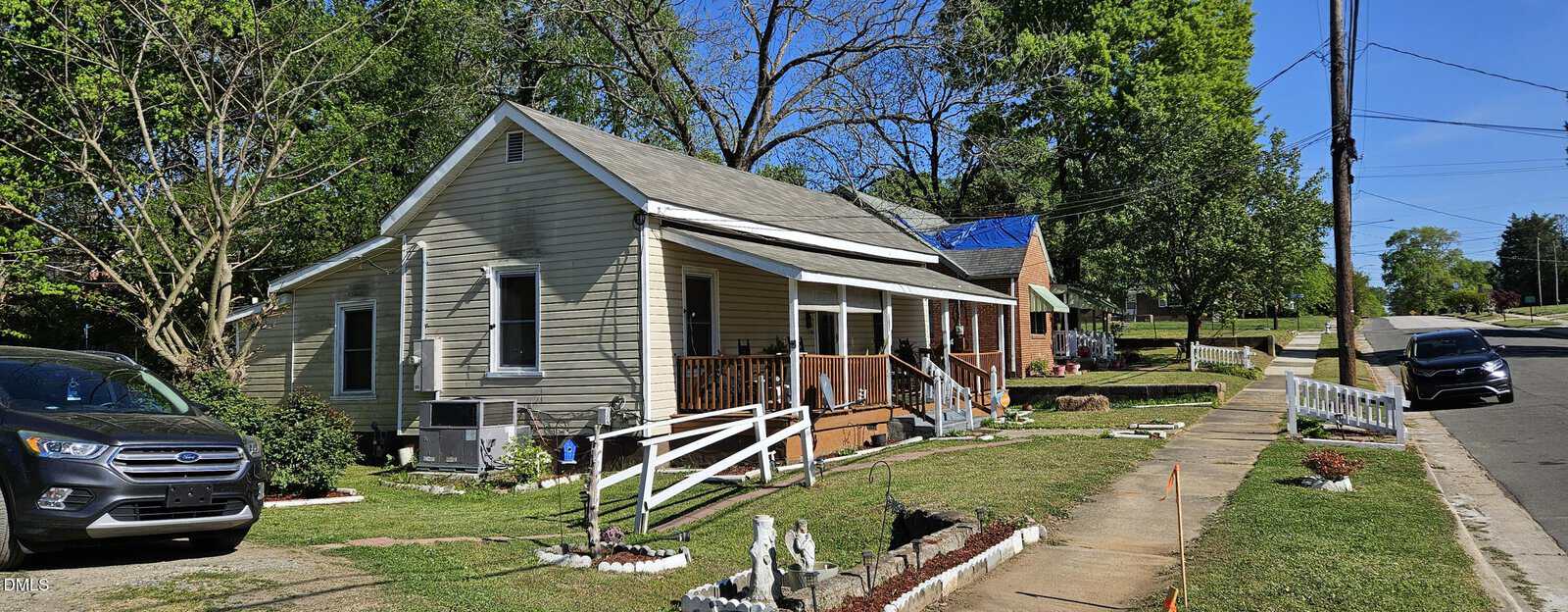 A beige single-story house with a front porch and ramp sits on a grassy corner lot. A black car is parked in front, with other houses, trees, and a sidewalk lining the street on a sunny day.