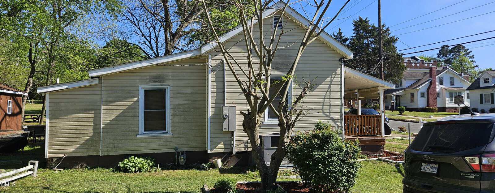 A beige single-story house with a covered side porch, a small tree and shrubs in the front yard, a black car parked on the driveway, and other houses and trees visible in the background on a sunny day.