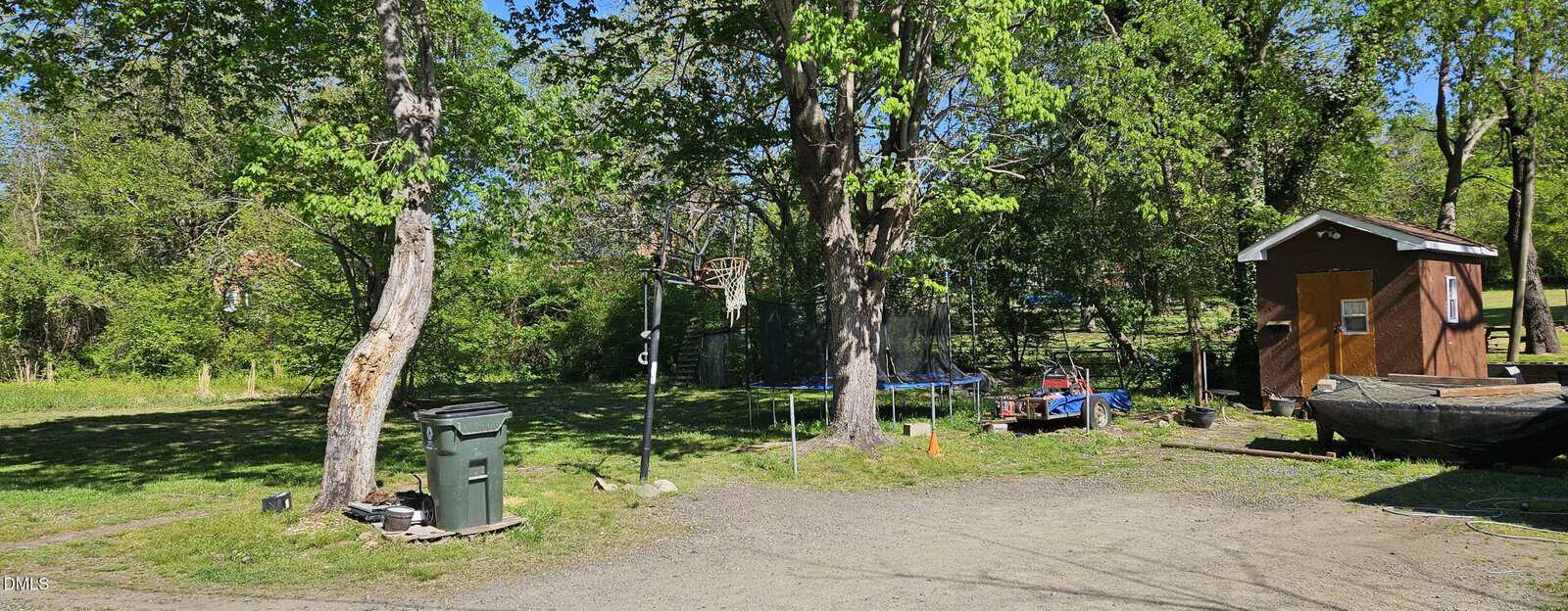 A sunny backyard with green trees, a trampoline, a small brown shed, a trash bin, boats, and gravel ground. Lush greenery is in the background.