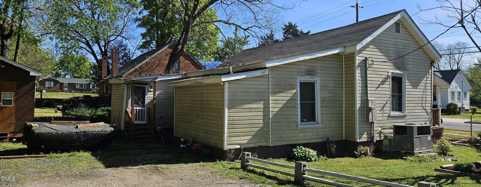 A small, light-yellow house with a sloped roof sits on a corner lot. There is an attached porch and wooden steps leading to a door, with trees and neighboring houses visible in the background.