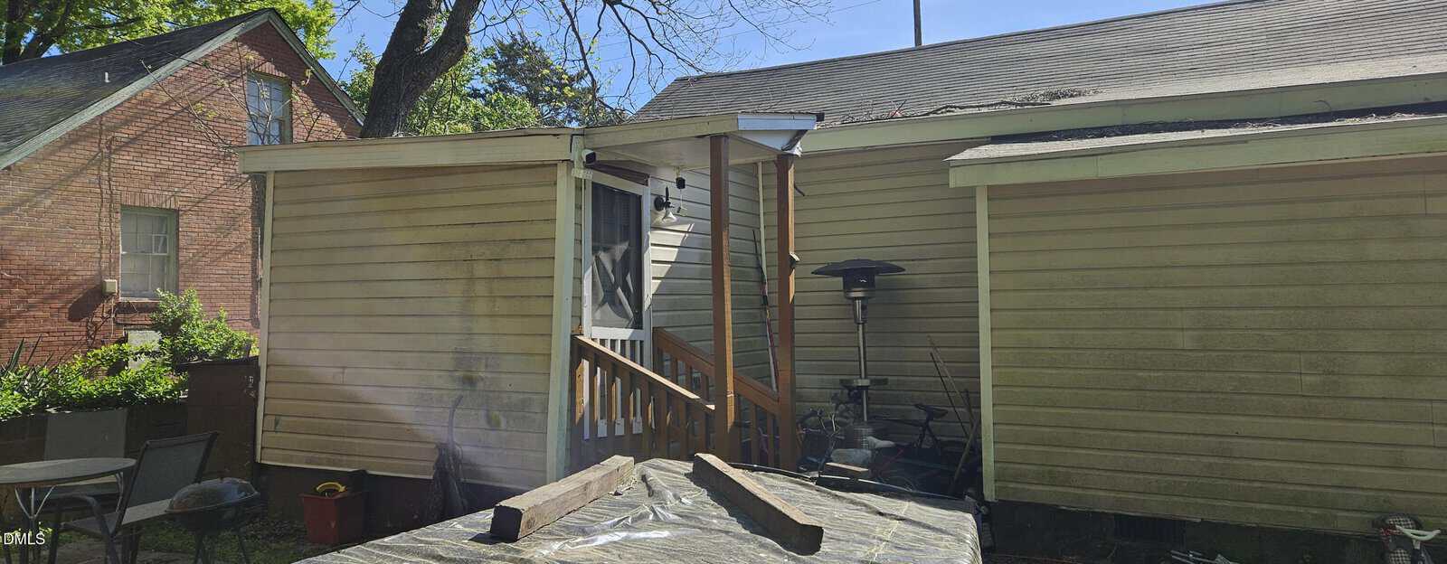 A small covered back porch with wooden steps leads to the entrance of a tan house. Patio items, a heater, and a table are nearby. A brick house and greenery are visible in the background under a clear sky.