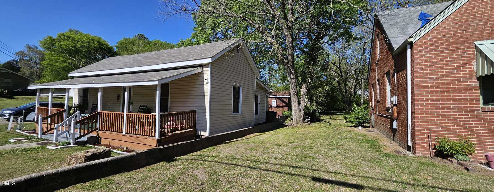 A beige house with a front porch sits beside a red brick house on a grassy yard with trees and blue sky in the background.