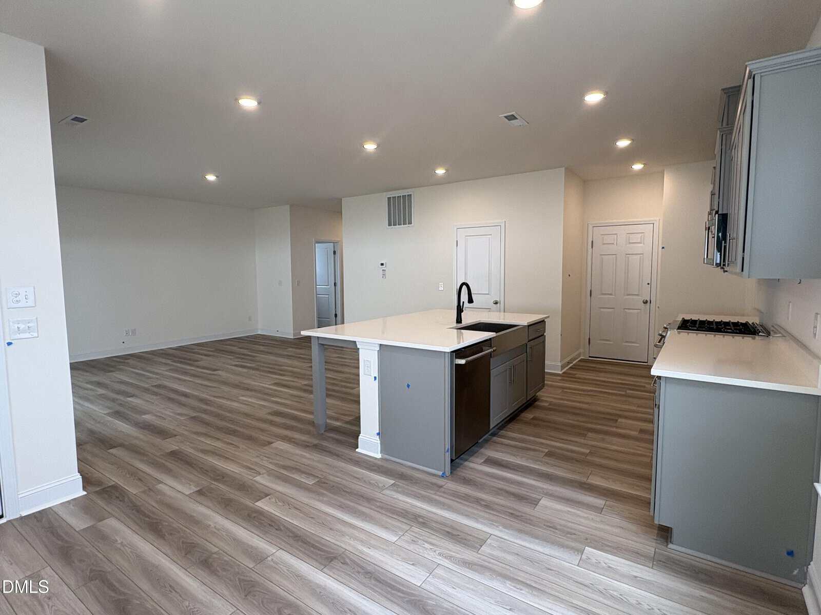 Modern open-concept kitchen and living area with light wood floors, white walls, recessed lighting, and a large island with a sink. The kitchen features gray cabinets and stainless steel appliances.