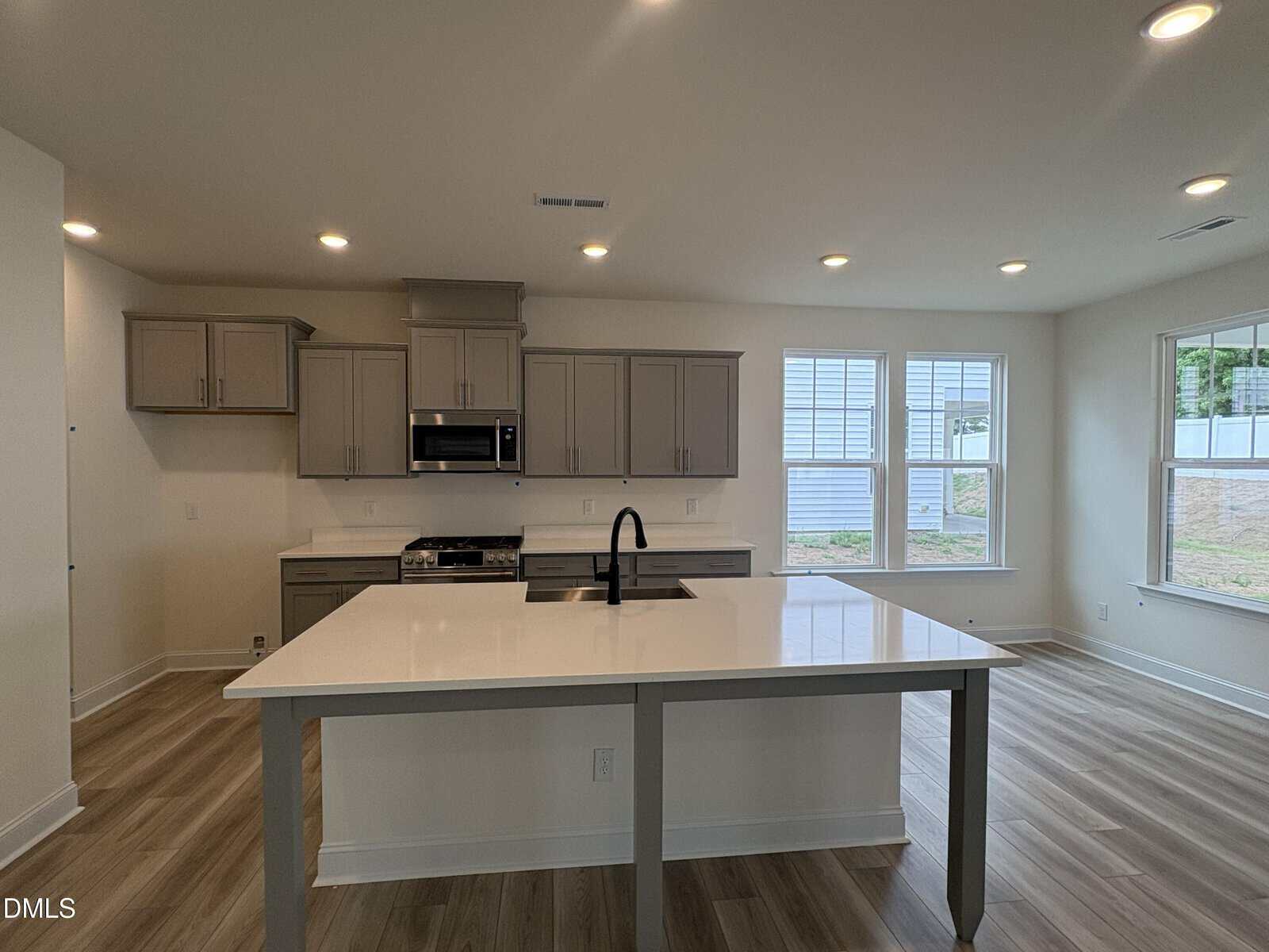 Modern kitchen with gray cabinets, stainless steel appliances, white island with built-in sink, wood-style flooring, recessed lighting, and large windows letting in natural light.