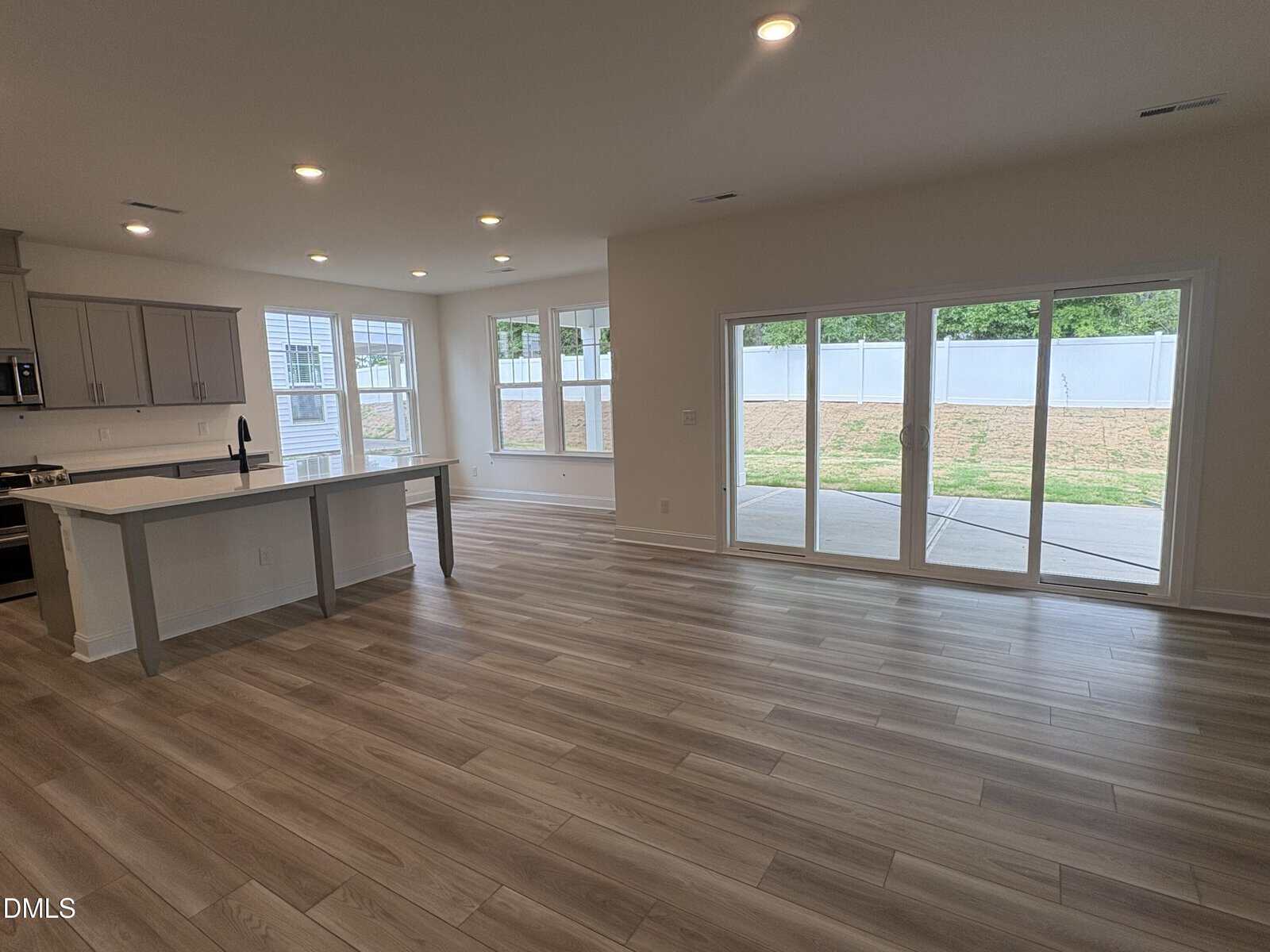 Spacious open-plan kitchen and living area with light wood flooring, large windows, and glass doors leading to a patio and fenced backyard. Kitchen features gray cabinets and a central island with a sink.