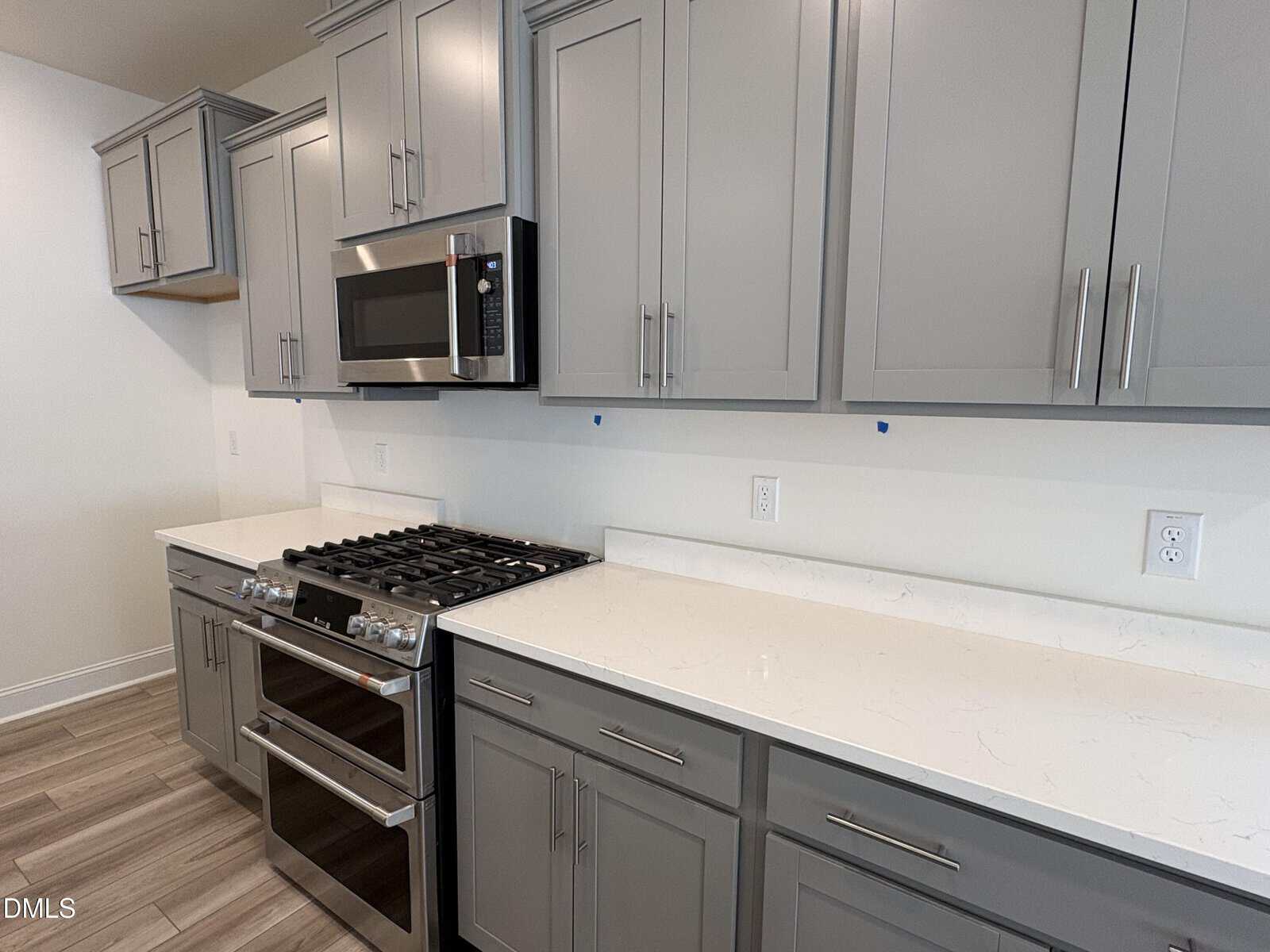 Modern kitchen with gray cabinets, stainless steel gas stove and oven, built-in microwave, white marble countertops, and light wood flooring. The space looks clean and uncluttered.