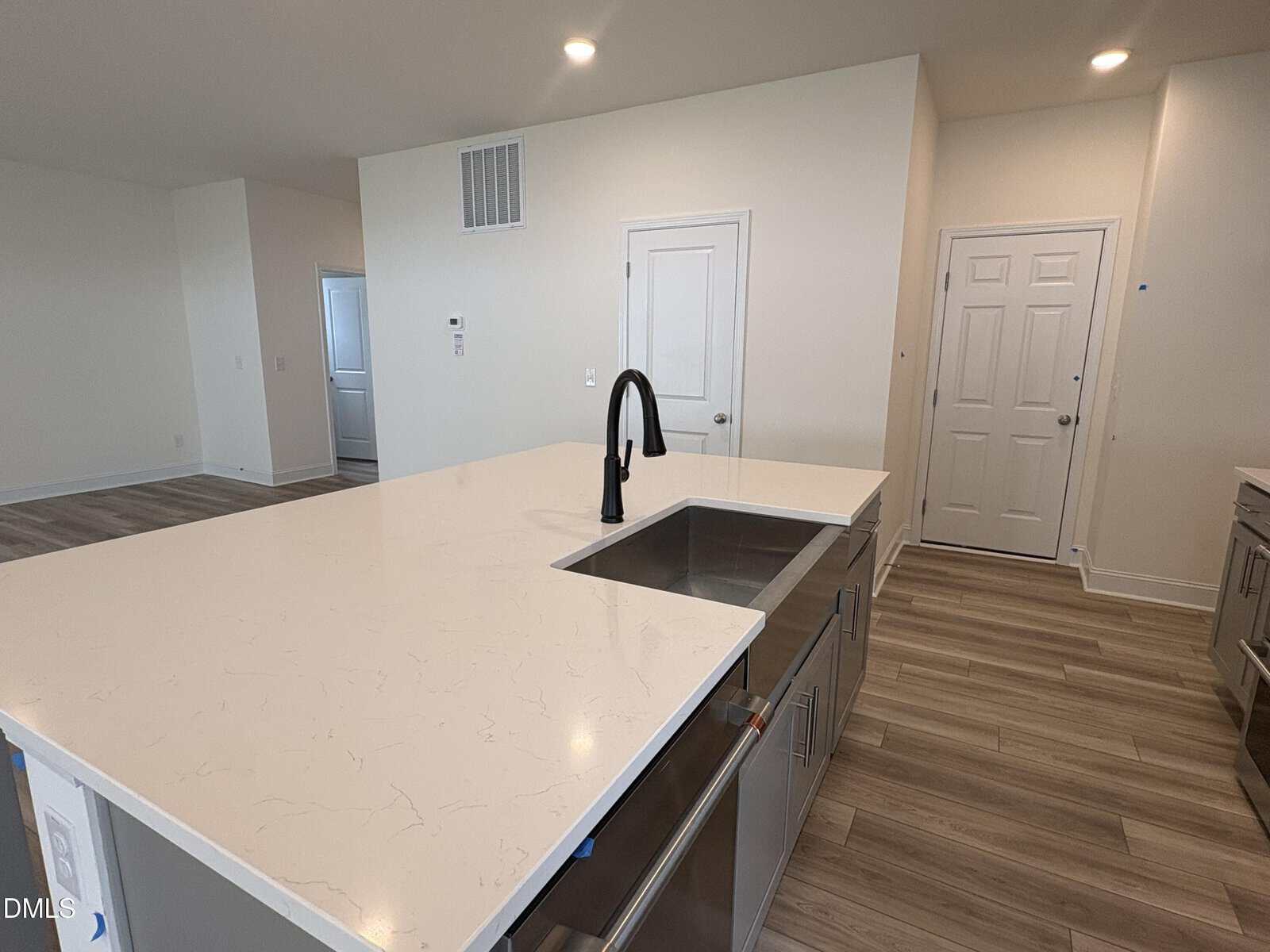 Modern kitchen with a large white island, black faucet, stainless steel sink, wood-style flooring, and white walls. Two closed white doors are visible in the background.
