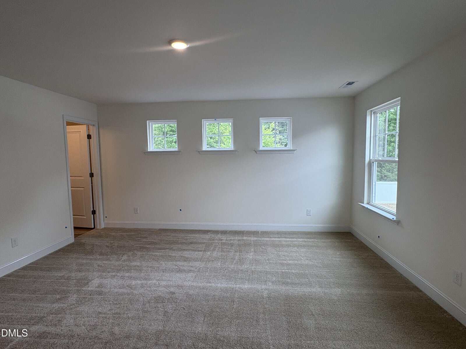 Empty carpeted room with three small windows on one wall, one large window on the right, white walls, and a door to the left leading to another space. Ceiling light is turned on.