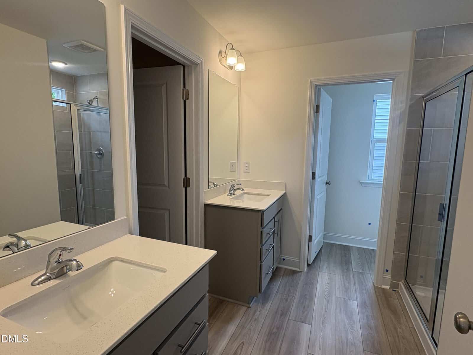 Modern bathroom with double sinks, separate mirrors, gray cabinets, wood-style flooring, and a glass shower enclosure. A closed door and a window are visible in the background.