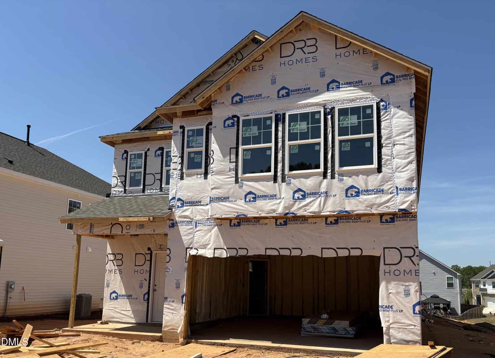 Two-story house under construction, exterior covered in white weather wrap branded with “DRB Homes.” Windows are installed; garage and front door are open. Construction materials are scattered on the dirt driveway.