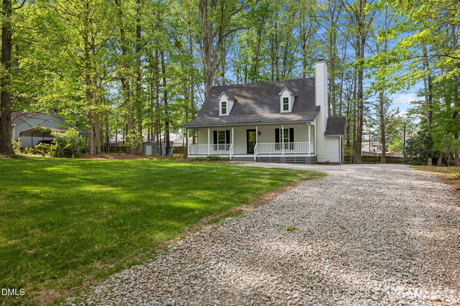 A white two-story house with a front porch and dormer windows, surrounded by tall trees, sits at 157 Mill Creek Drive in Youngsville. A gravel driveway leads up to the house through a green yard under a blue sky.
