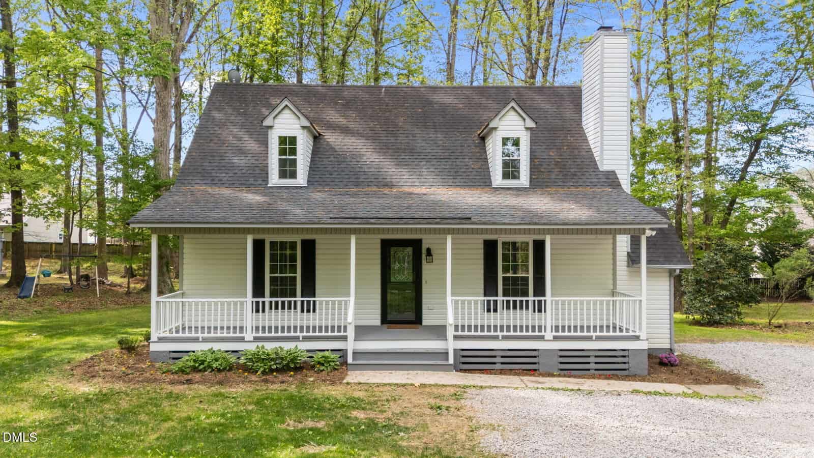A charming white house with a front porch, black shutters, dormer windows, and a gravel driveway, surrounded by trees and greenery on a sunny day.
