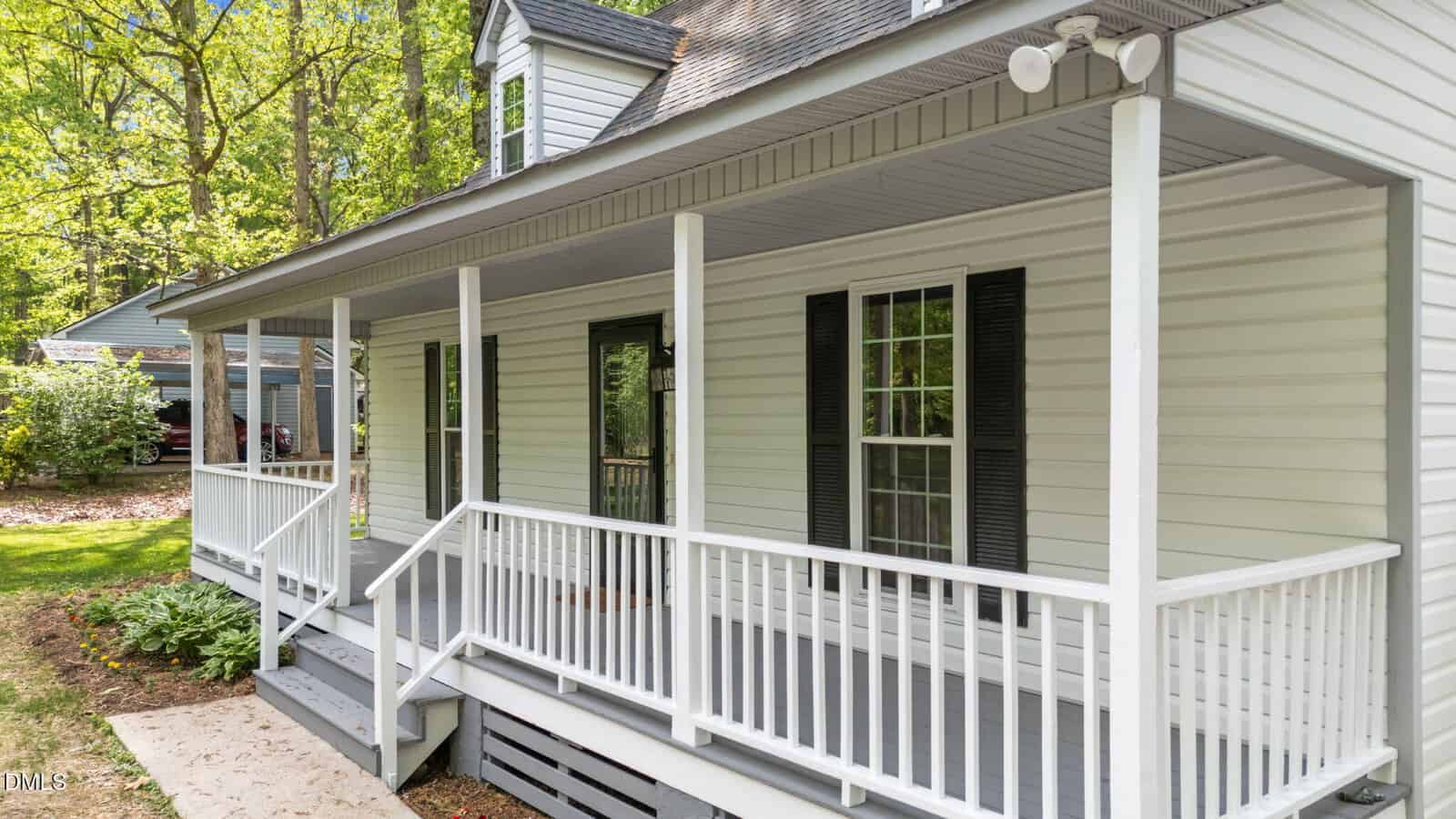 White house with a covered front porch, gray steps, black shutters, and surrounded by green trees and grass. The porch has white railings and columns, with a concrete path leading to the stairs.